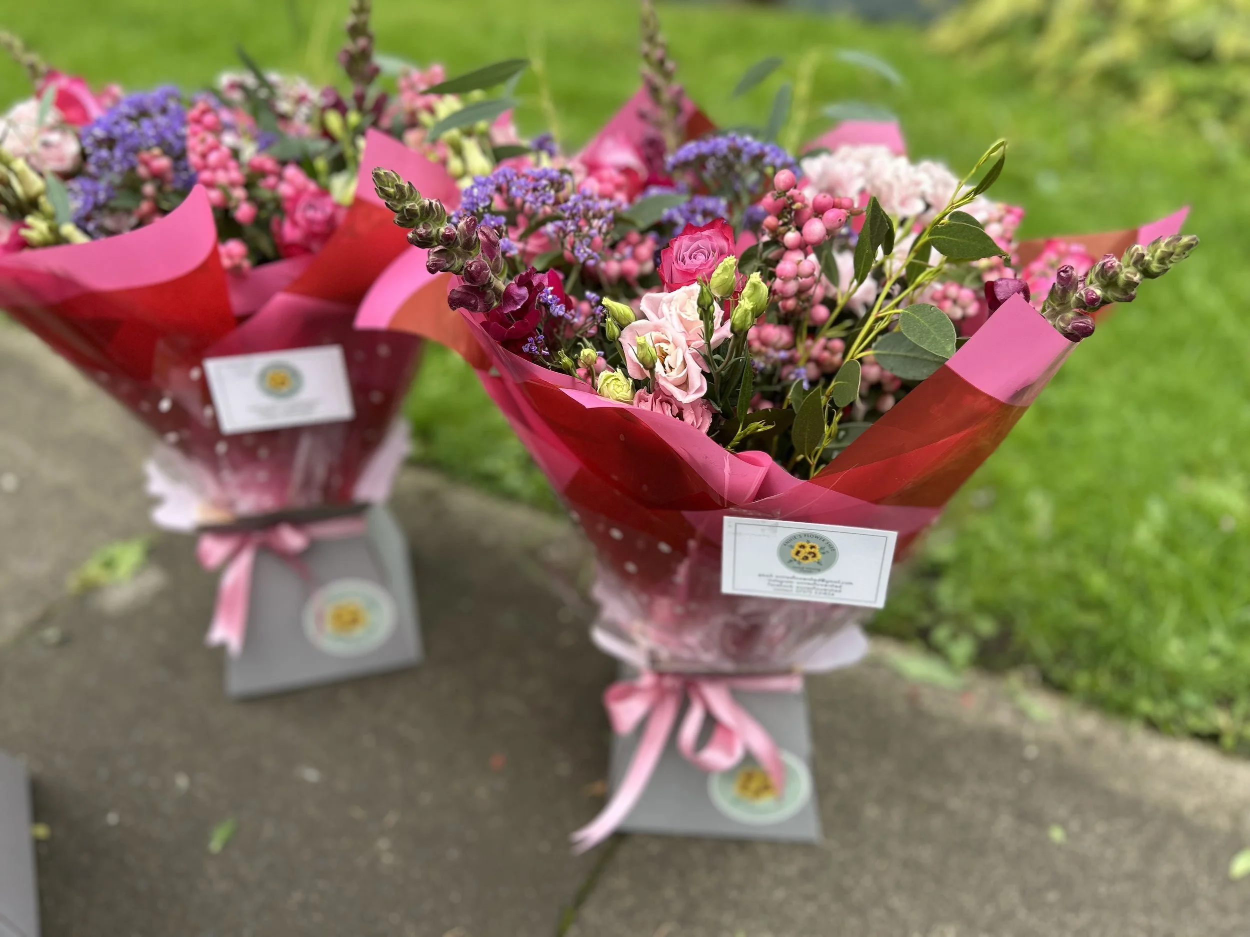 A bouquet of mixed flowers wrapped in pink and red paper with a label, displayed outdoors on a concrete surface, with a blurred green grass background.