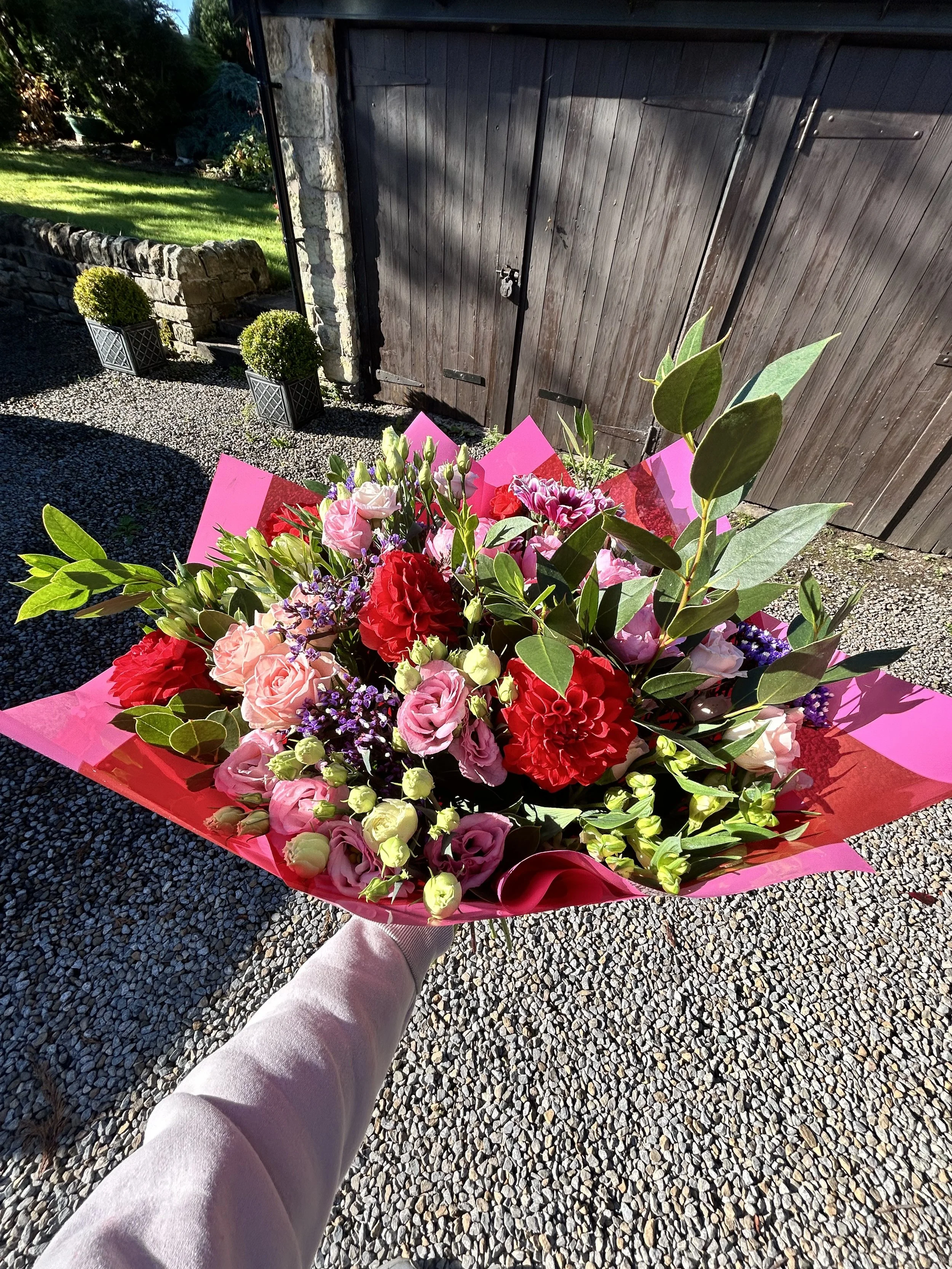 Colorful bouquet of pink, red, and purple flowers with green leaves, wrapped in pink paper, being held by a person wearing light-colored pants outdoors near a wooden shed.