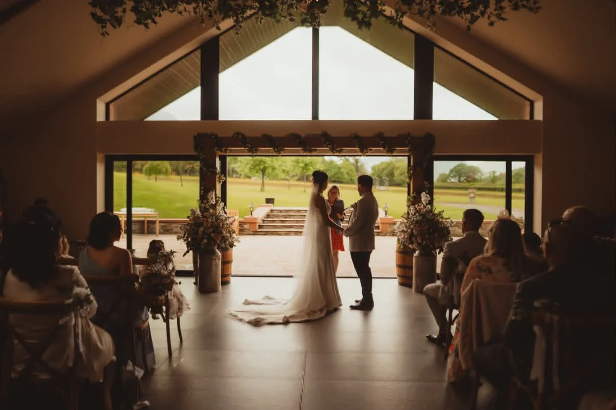 A bride and groom exchanging vows during their wedding ceremony inside a hall with large windows overlooking a green outdoor landscape.