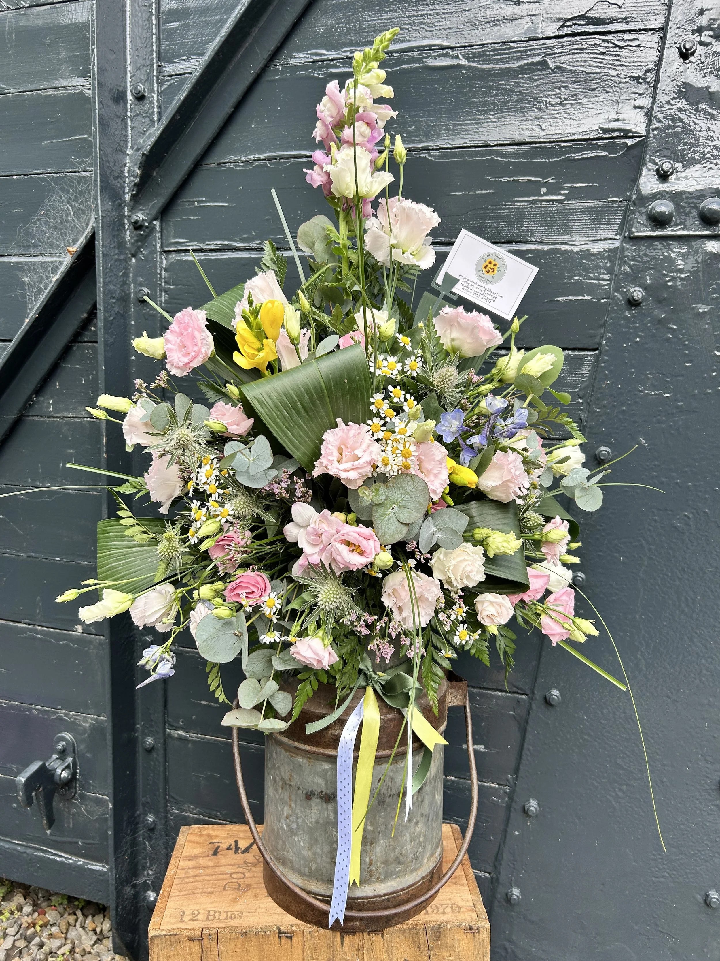 A floral arrangement in a rustic metal bucket with pink, white, yellow, purple, and green flowers and foliage, placed on a wooden box against a black weathered wooden wall.