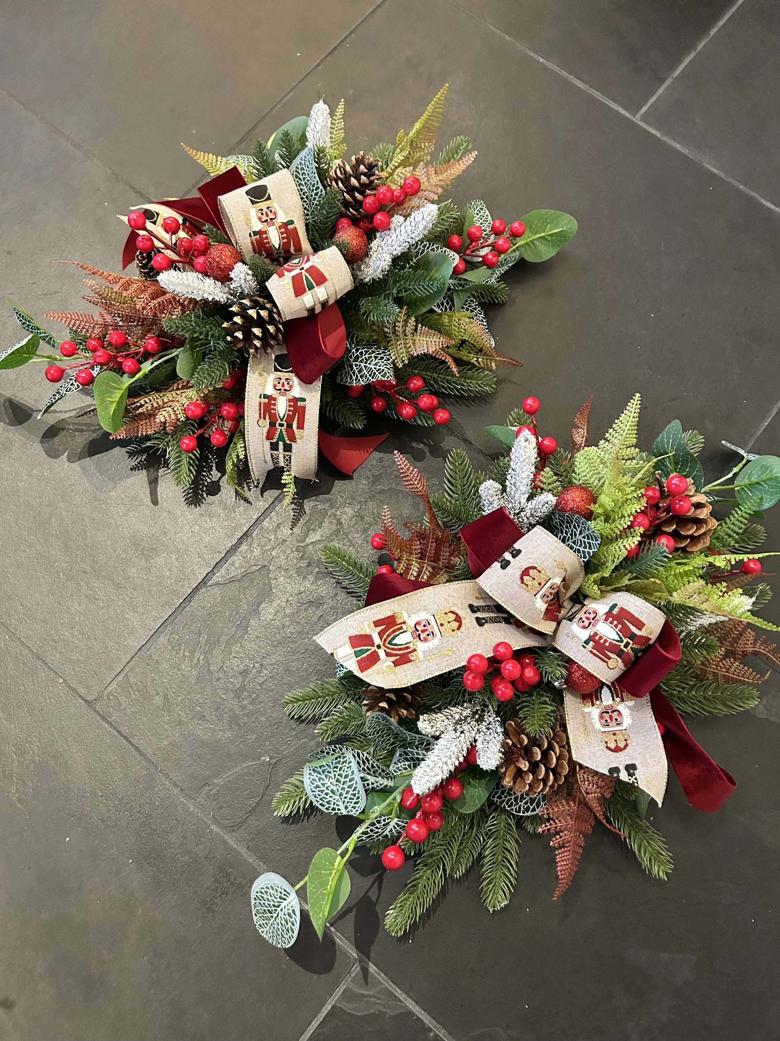 Two Christmas wreaths decorated with pinecones, red berries, and holiday ribbons, placed on a dark tiled floor.