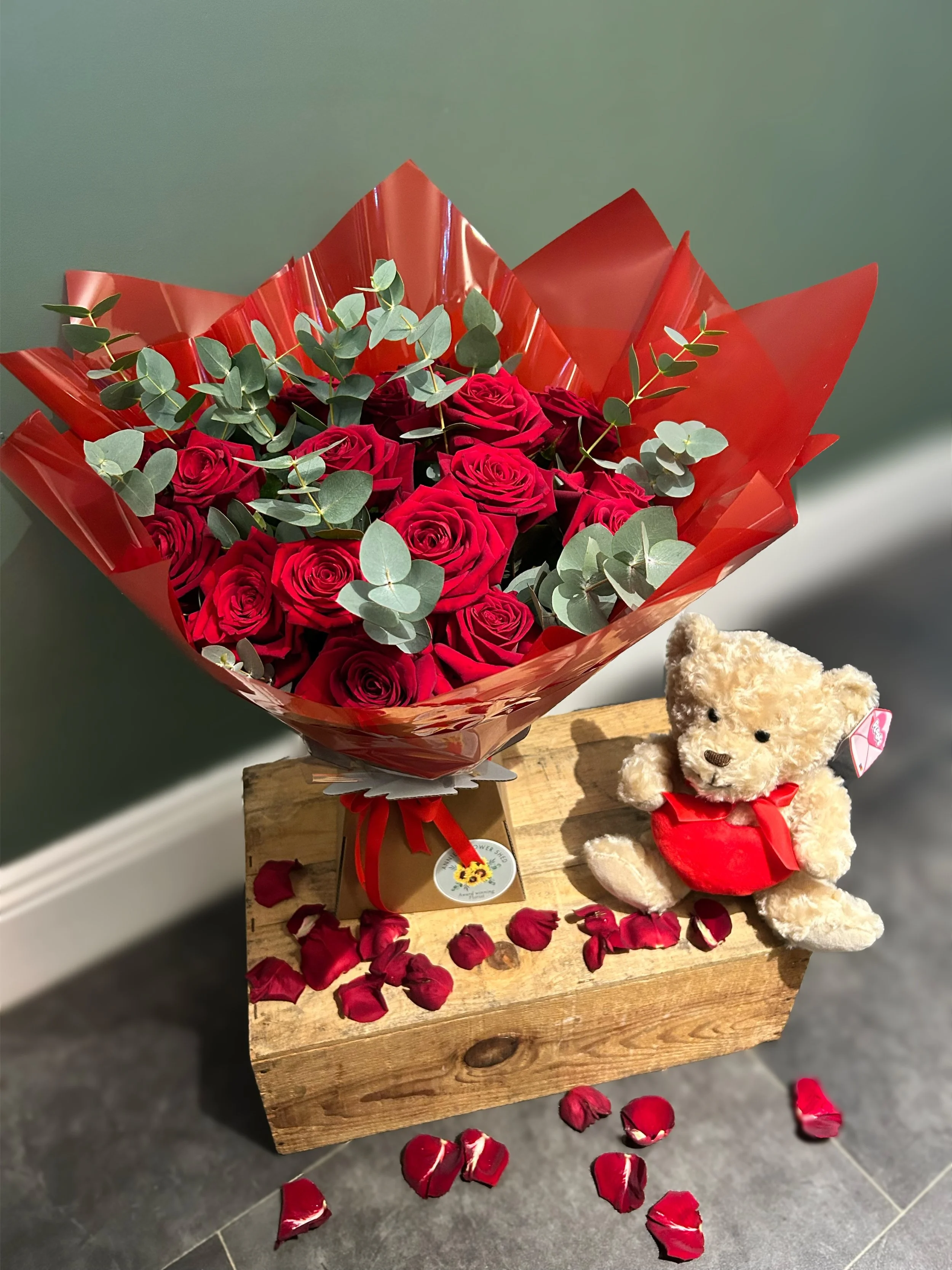 A bouquet of red roses with eucalyptus leaves wrapped in red paper, placed on a wooden table with scattered rose petals, and a small teddy bear with a red dress, all against a green wall background.