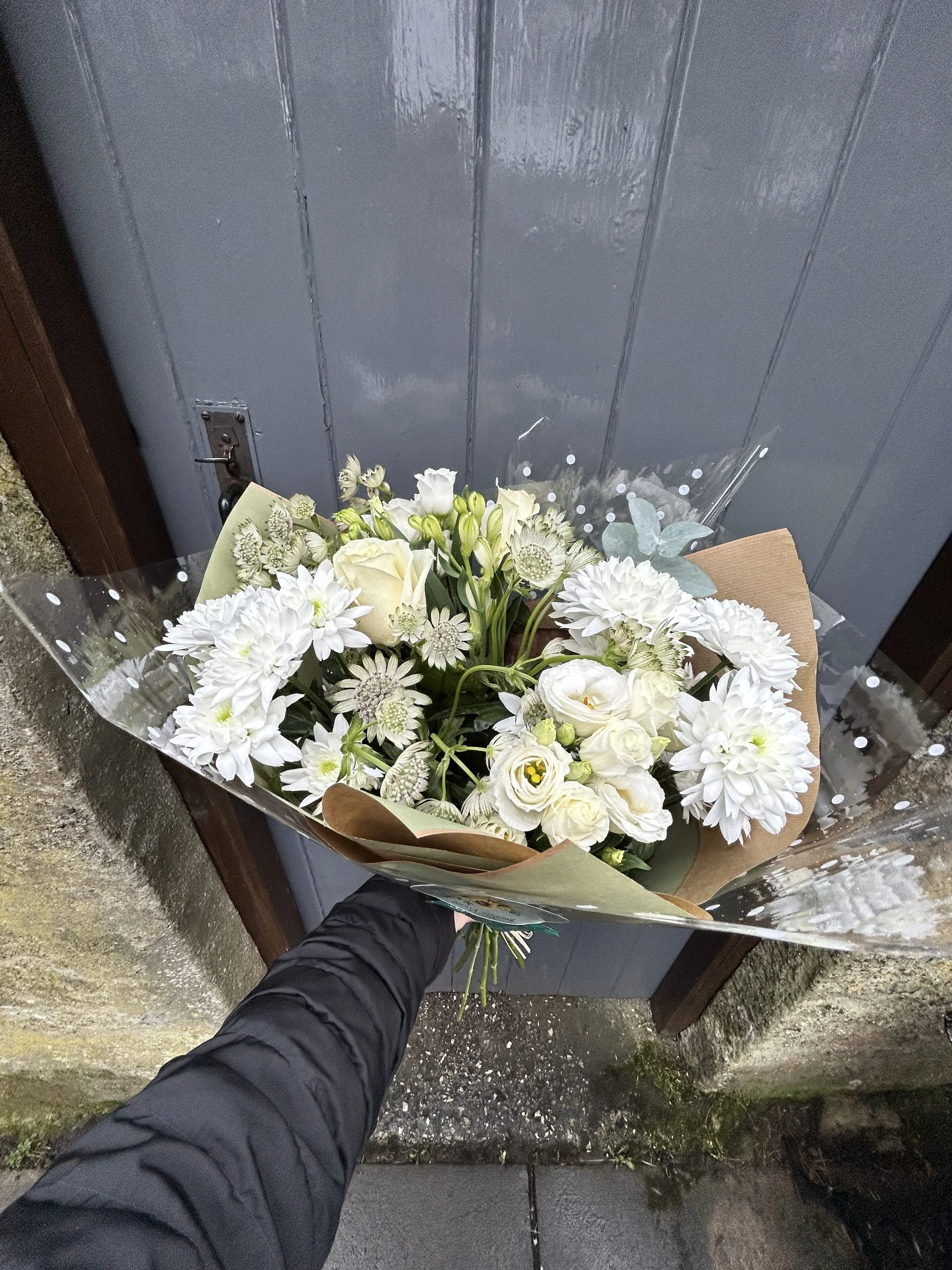 A person holding a bouquet of white flowers, standing outdoors near a gray door.