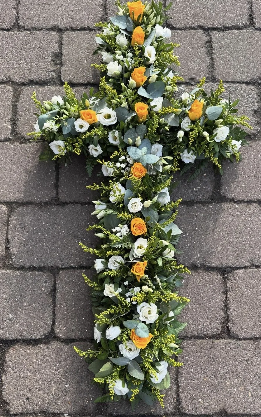 Flower arrangement shaped like a cross with white and yellow flowers, green leaves, and eucalyptus, placed on a brick pavement.