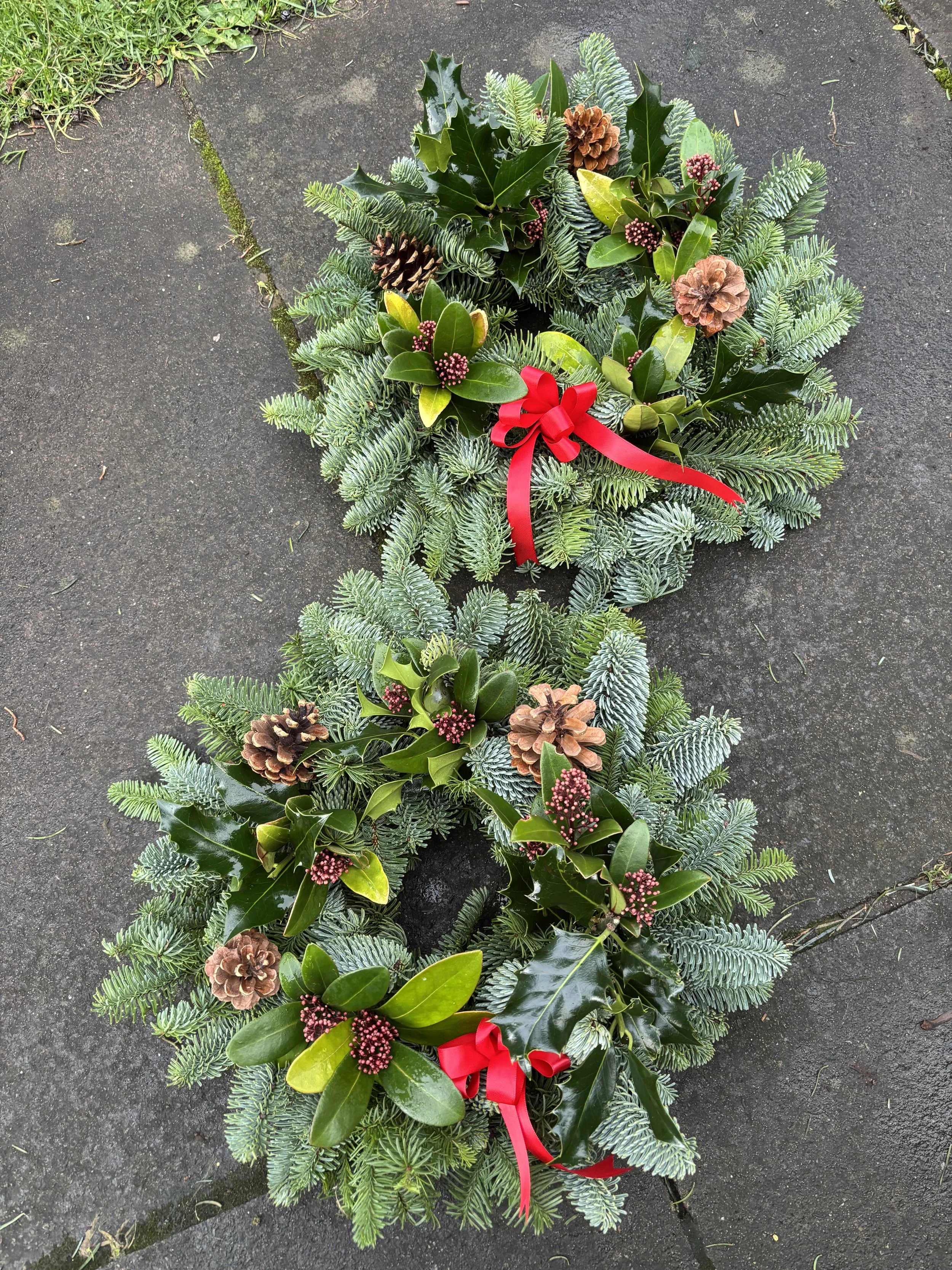 A Christmas wreath with pine, holly, pine cones, small red berries, and a red bow, placed on a dark concrete surface.