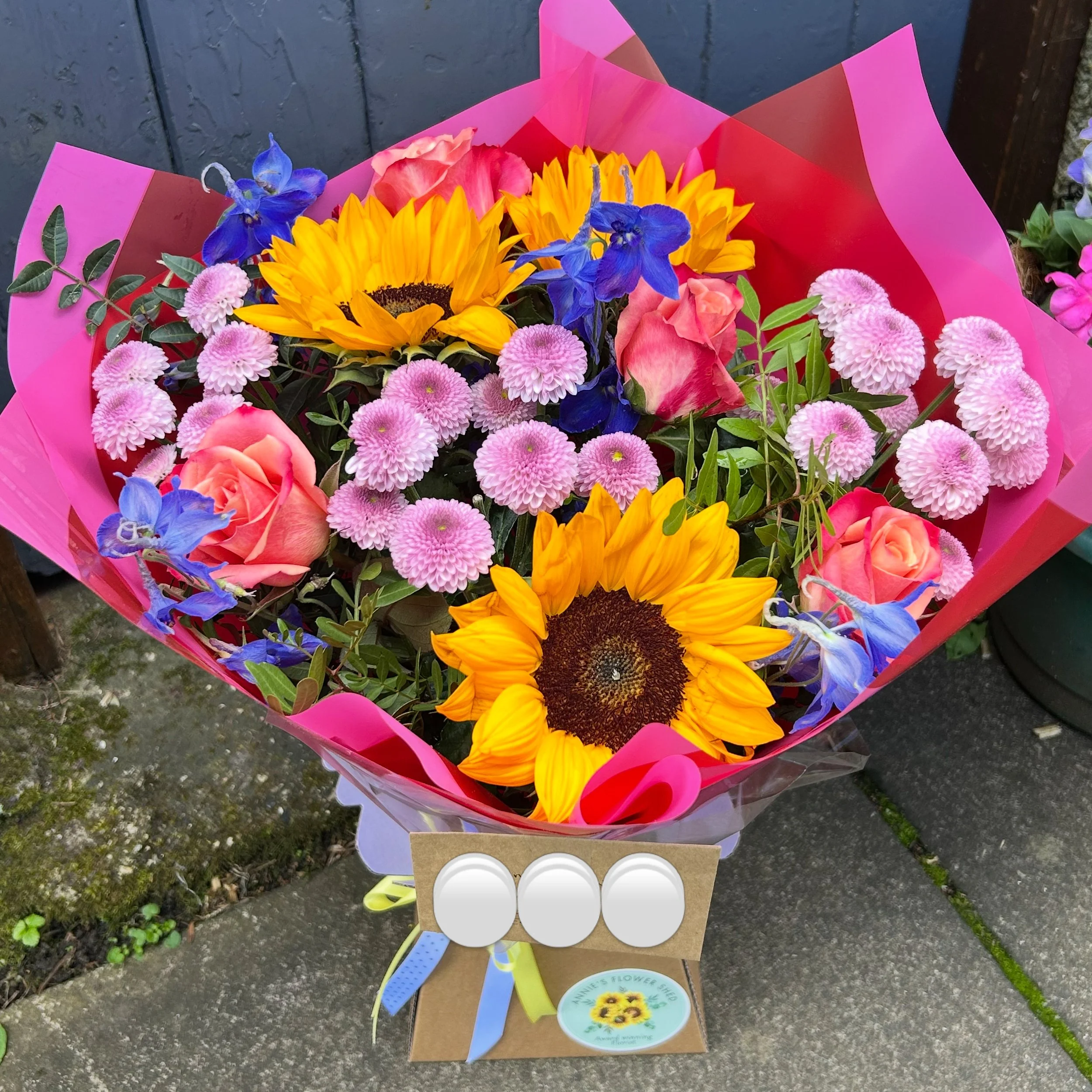 A colorful bouquet of mixed flowers wrapped in pink and red paper, including sunflowers, pink roses, blue irises, and pink chrysanthemums, with a flower shop sticker on the wrapping.
