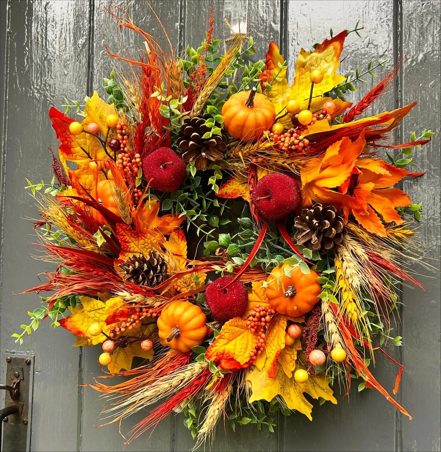 Autumn-themed wreath with small pumpkins, artificial red apples, pinecones, yellow and orange leaves, and wheat stalks hanging on a gray door.