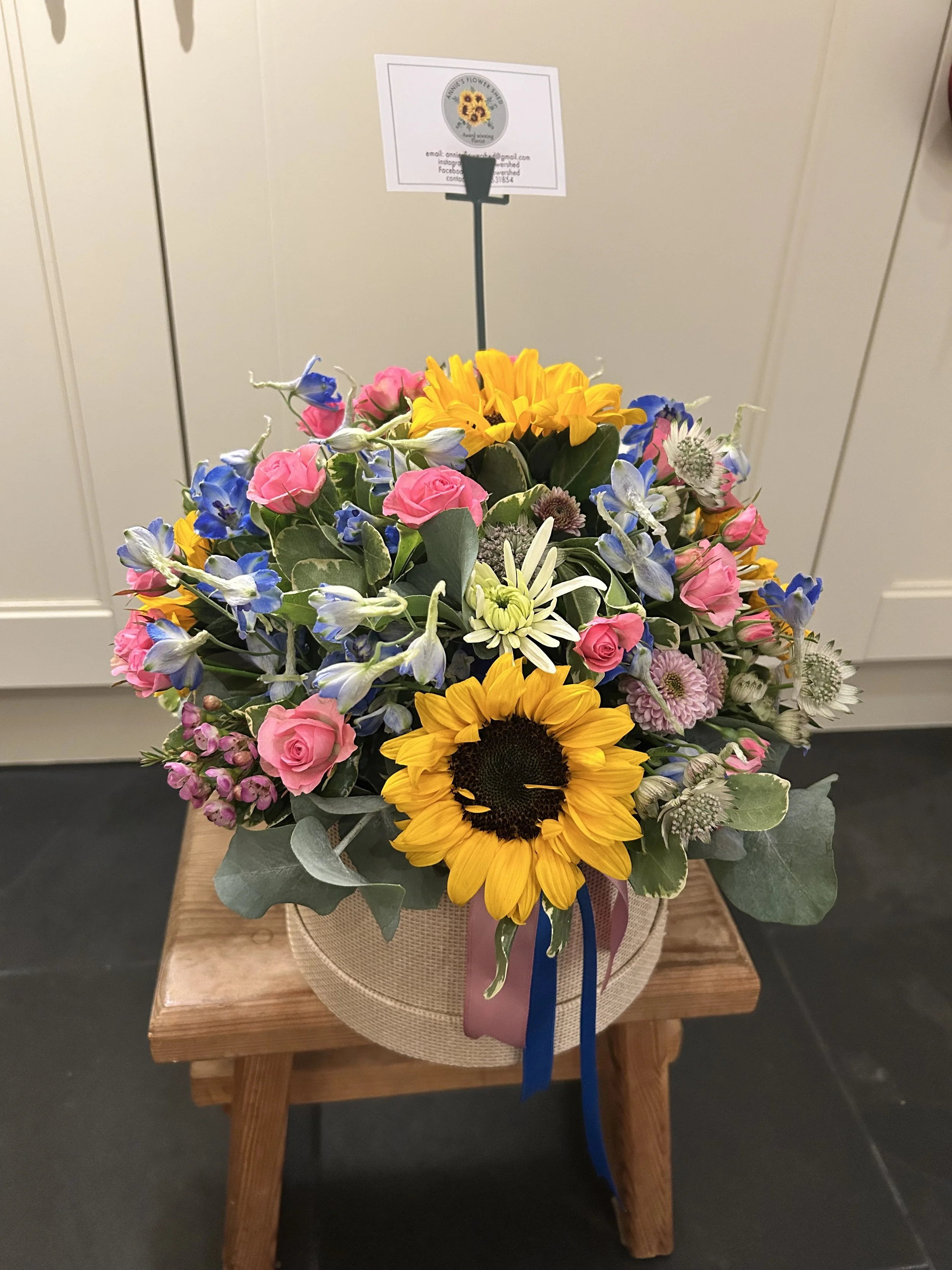 Mixed flower bouquet with sunflowers, pink roses, blue delphiniums, and other flowers on a wooden table with a beige wrapping and ribbon.