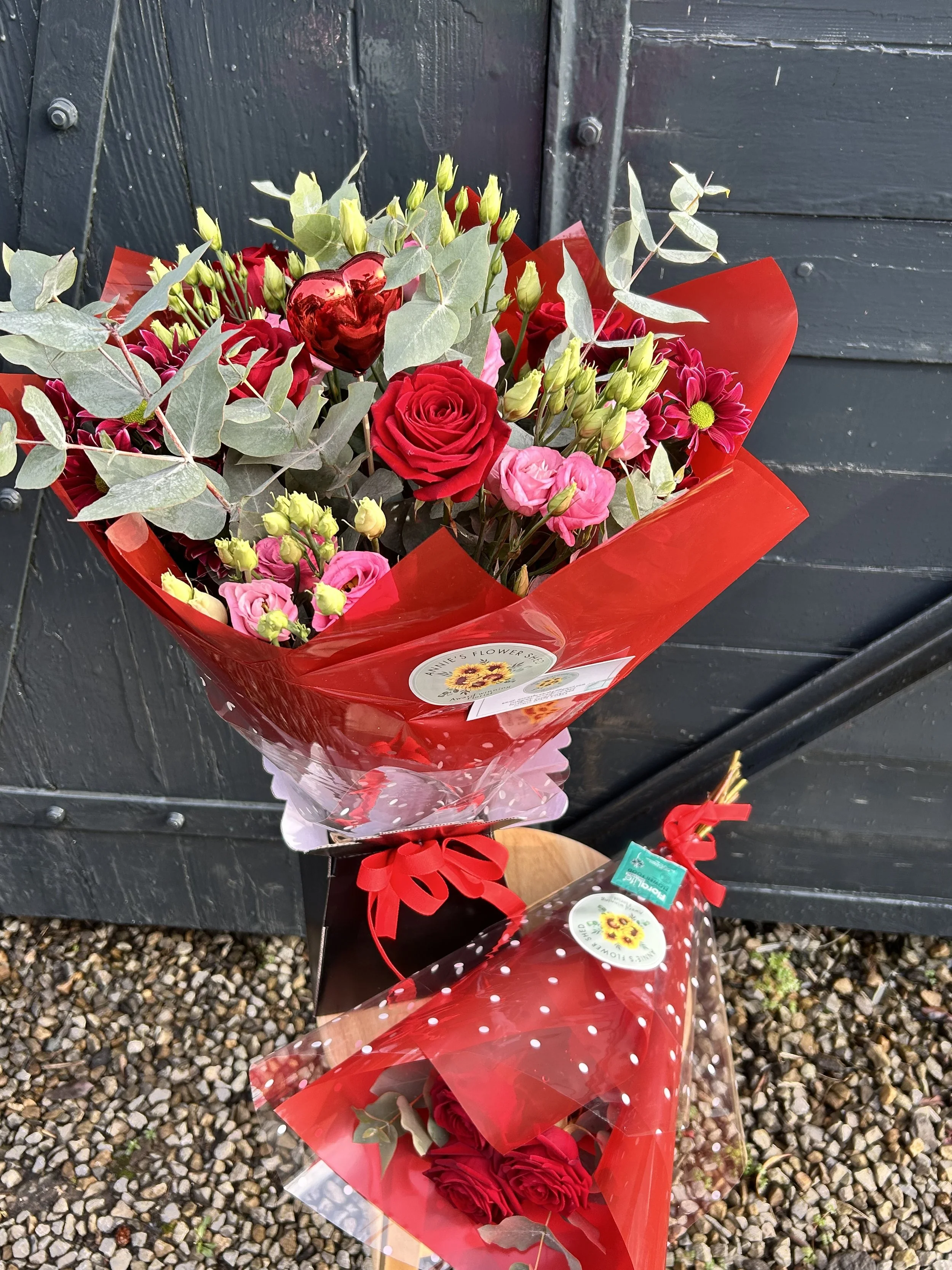 A bouquet of flowers wrapped in red paper, containing red roses, light pink roses, green buds, eucalyptus leaves, and a shiny red heart-shaped decoration, placed against a dark wooden background.