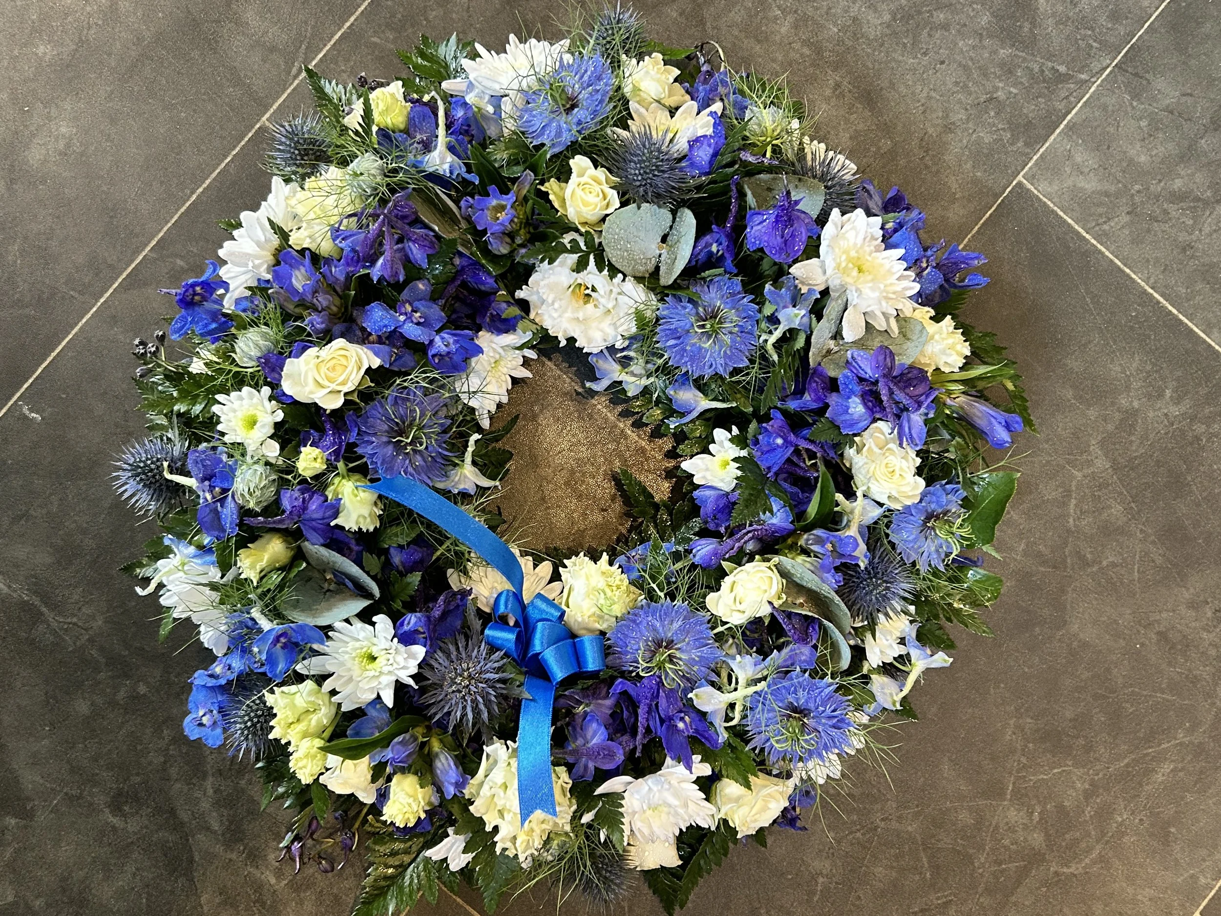 A floral arrangement in the shape of a heart featuring predominantly blue and white flowers, accented with green leaves and a blue ribbon, placed on a tiled gray floor.