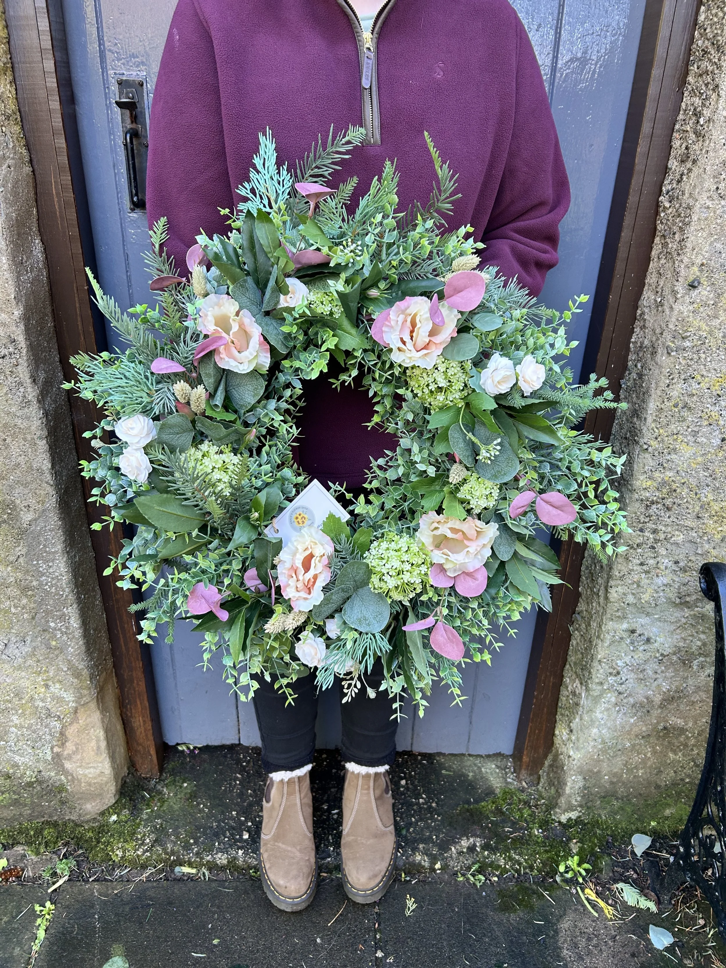 Person holding a large floral wreath standing in front of a blue door with a stone wall on the sides. The person is wearing a purple fleece jacket, black pants, and tan boots.