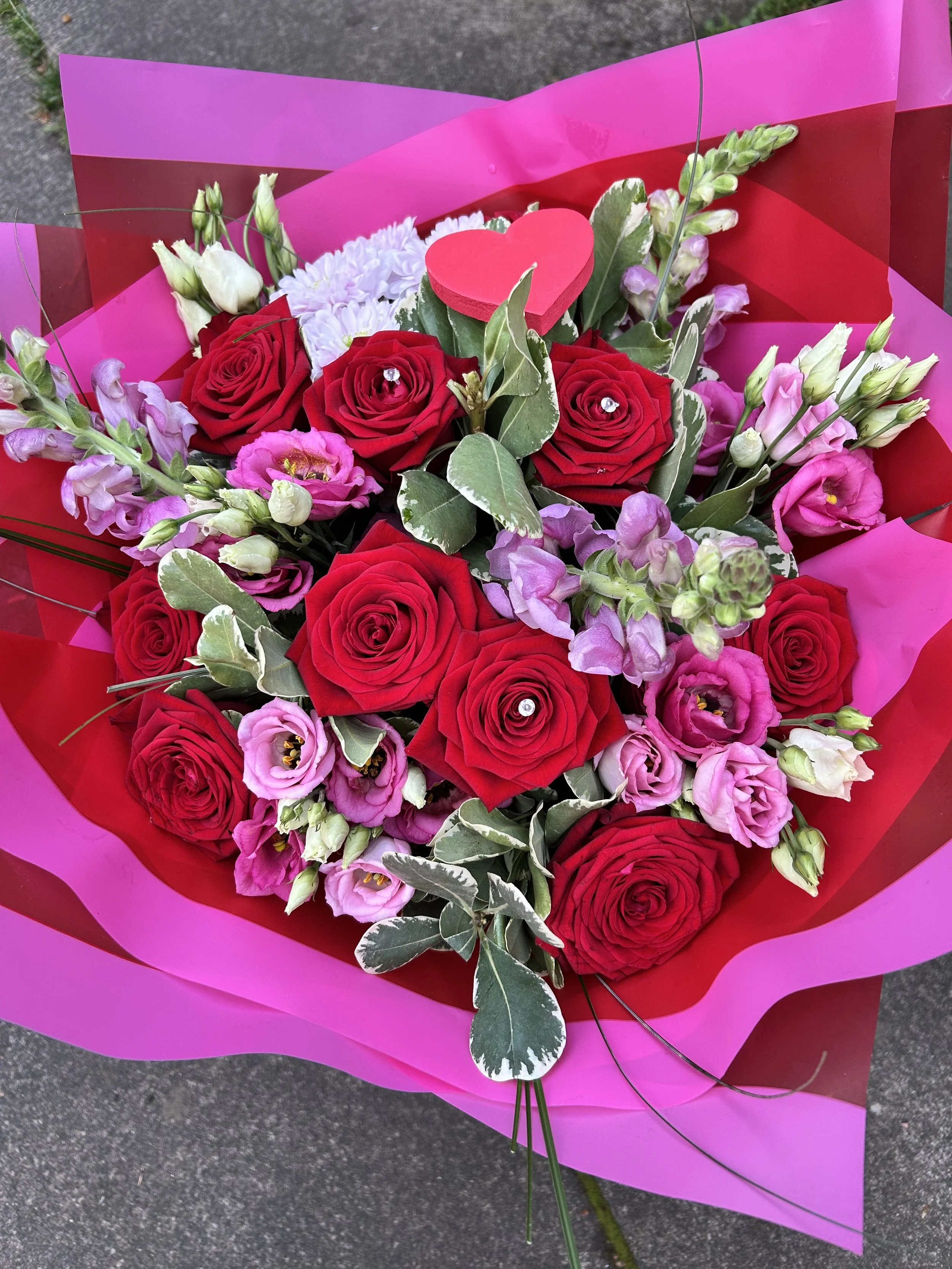 Bouquet of red roses, pink lisianthus, white chrysanthemums, and variegated leaves wrapped in pink and red paper with a small pink heart decoration.