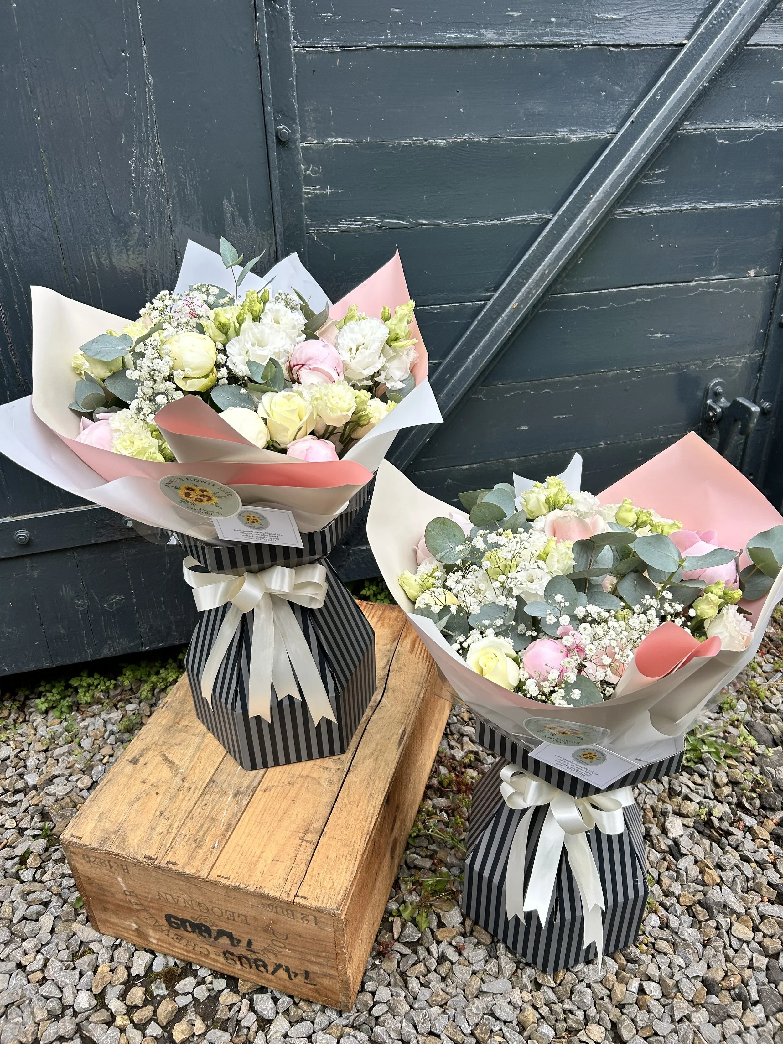 Two floral bouquets placed on a wooden platform with a dark wooden door in the background. The bouquets contain white, pink, and green flowers and are wrapped in decorative paper, tied with ribbons.