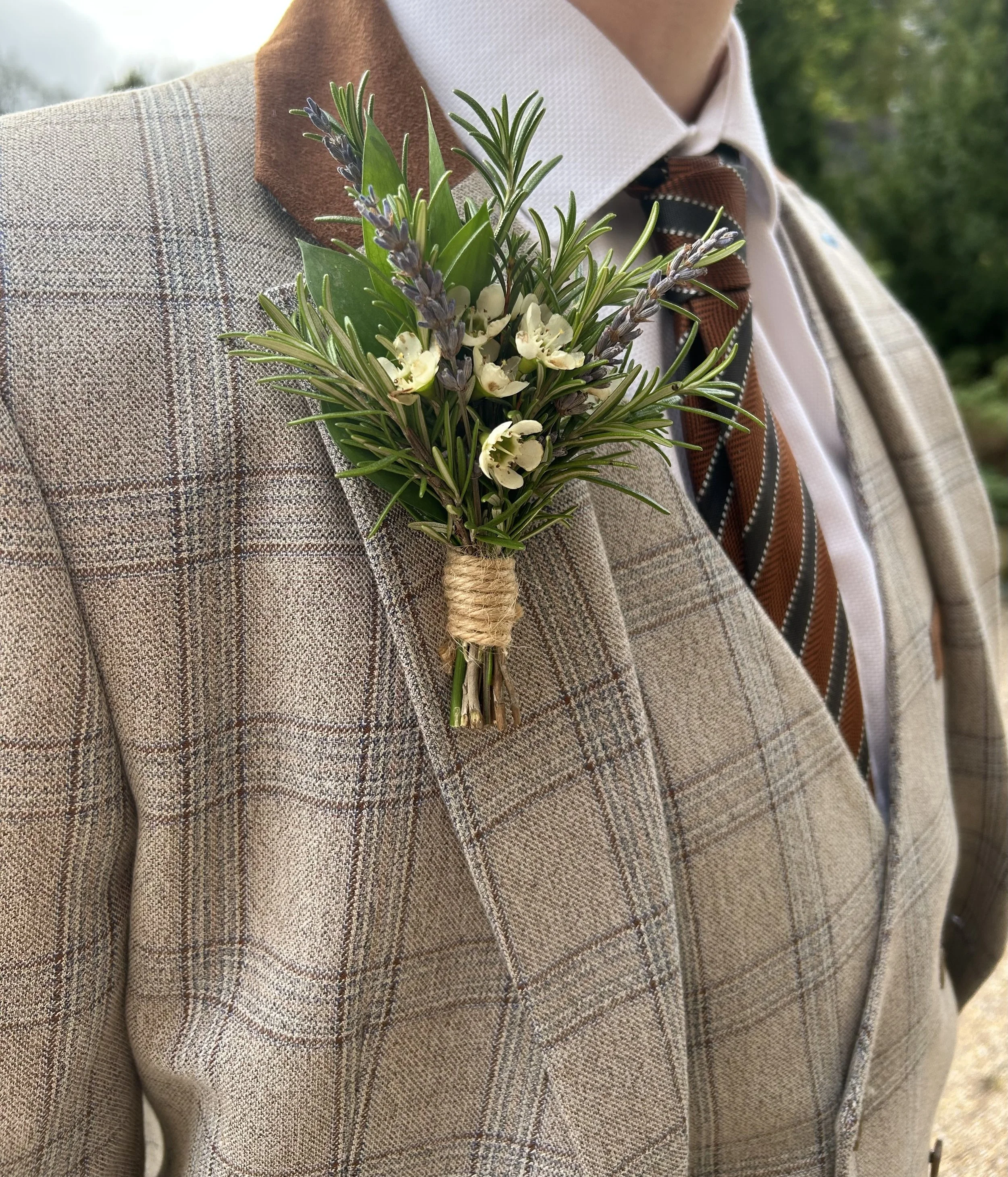 Close-up of a boutonniere with green leaves, lavender flowers, and white blossoms, worn on the lapel of a man's beige plaid suit jacket during a formal event.