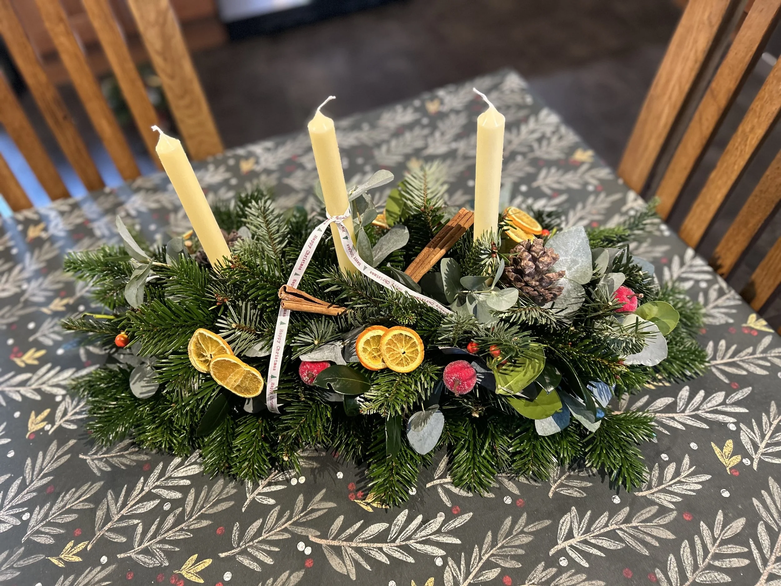 A holiday centerpiece with three unlit white candles surrounded by pine branches, dried orange slices, cinnamon sticks, pine cones, and decorative foliage on a festive tablecloth.
