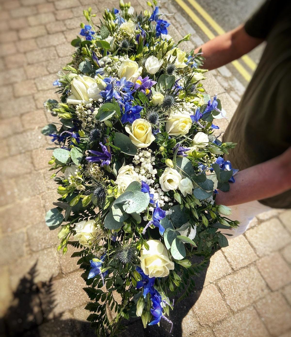 Person holding a large floral arrangement with white roses, purple and blue flowers, white baby's breath, and greenery, outdoors on a brick-paved surface.