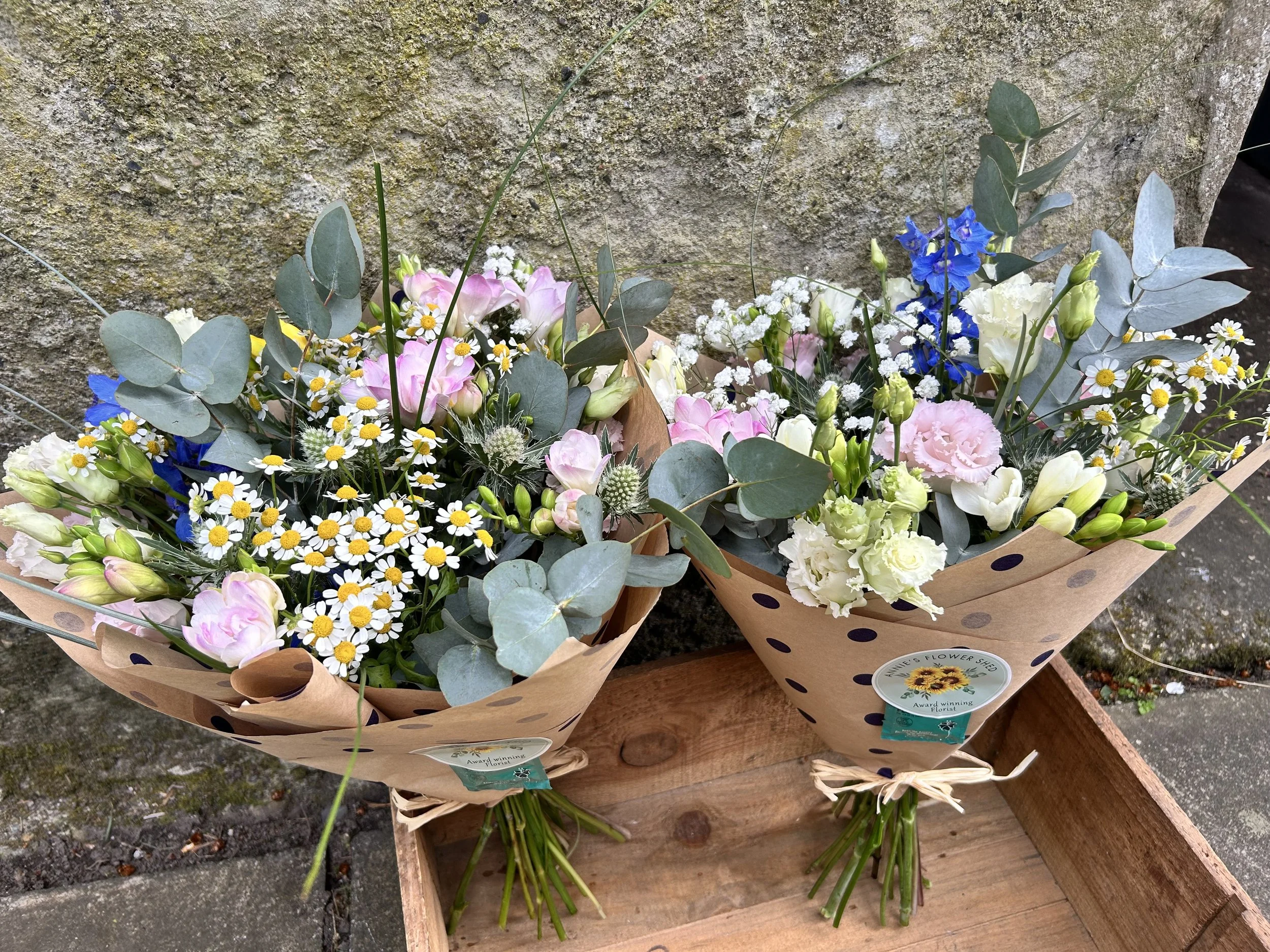 Two bouquets of mixed flowers including daisies, pink roses, blue irises, and eucalyptus on a wooden surface in front of a textured stone wall.