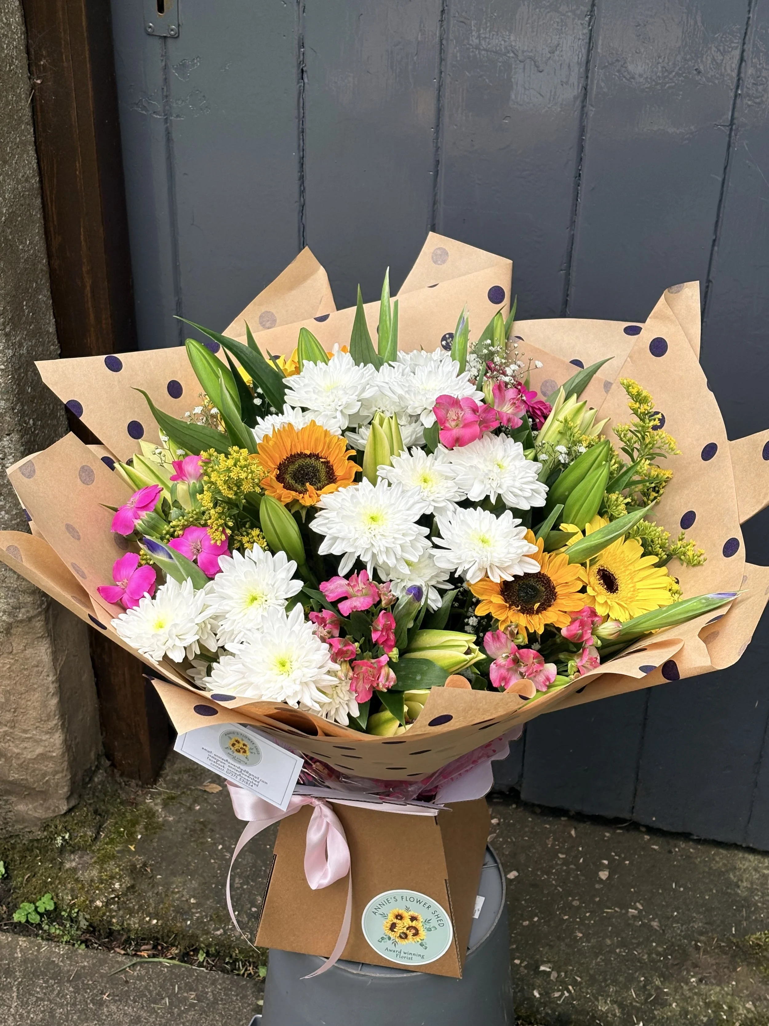 A colorful bouquet of flowers including white chrysanthemums, orange sunflowers, pink and purple flowers, and green foliage, wrapped in brown polka-dotted paper with a pink ribbon, placed on top of a trash bin against a dark blue wooden door and conc