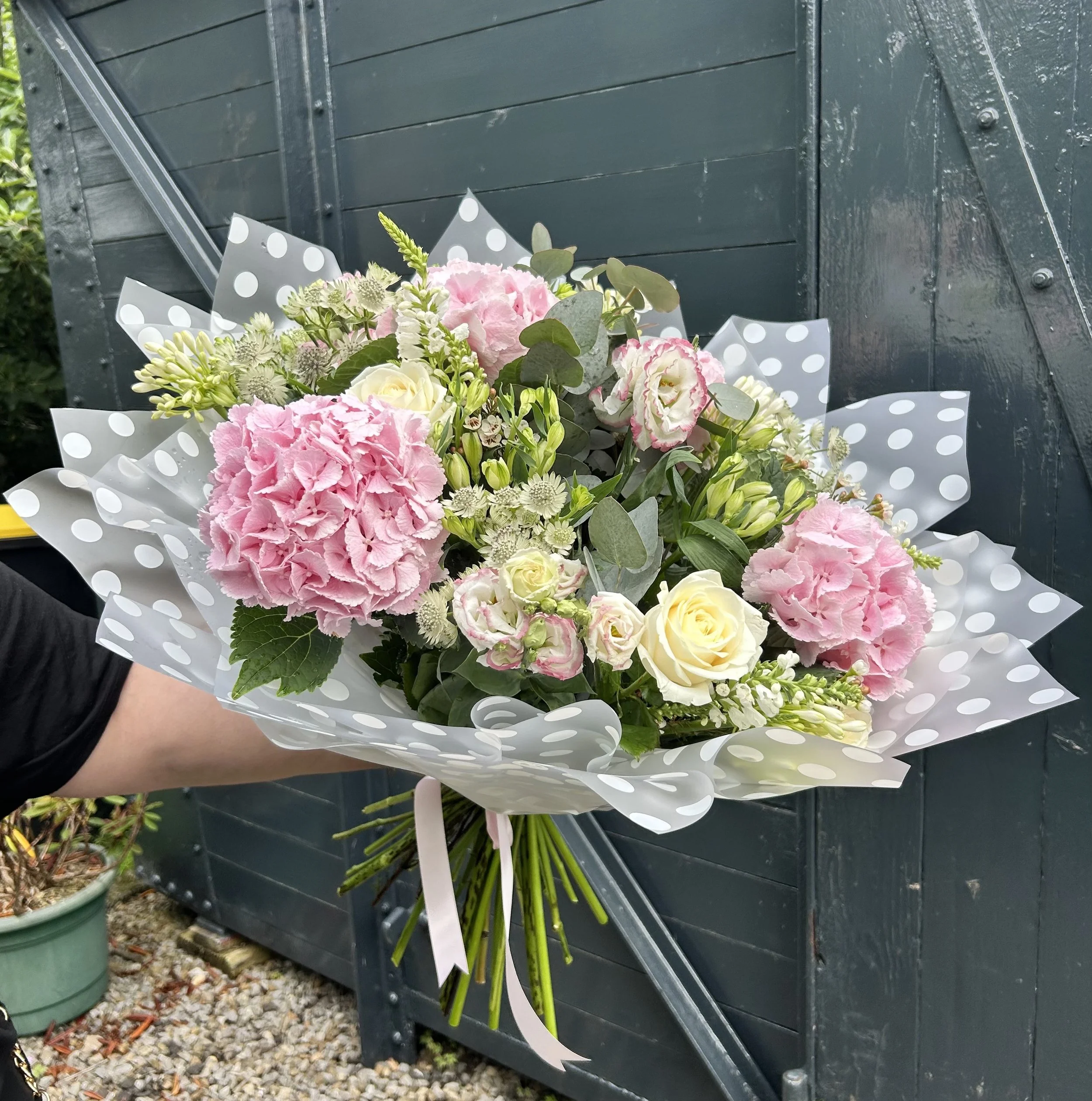 A bouquet of pink, white, and cream flowers with green leaves and wrapping paper leaning against a dark gray wooden wall with metal hardware.