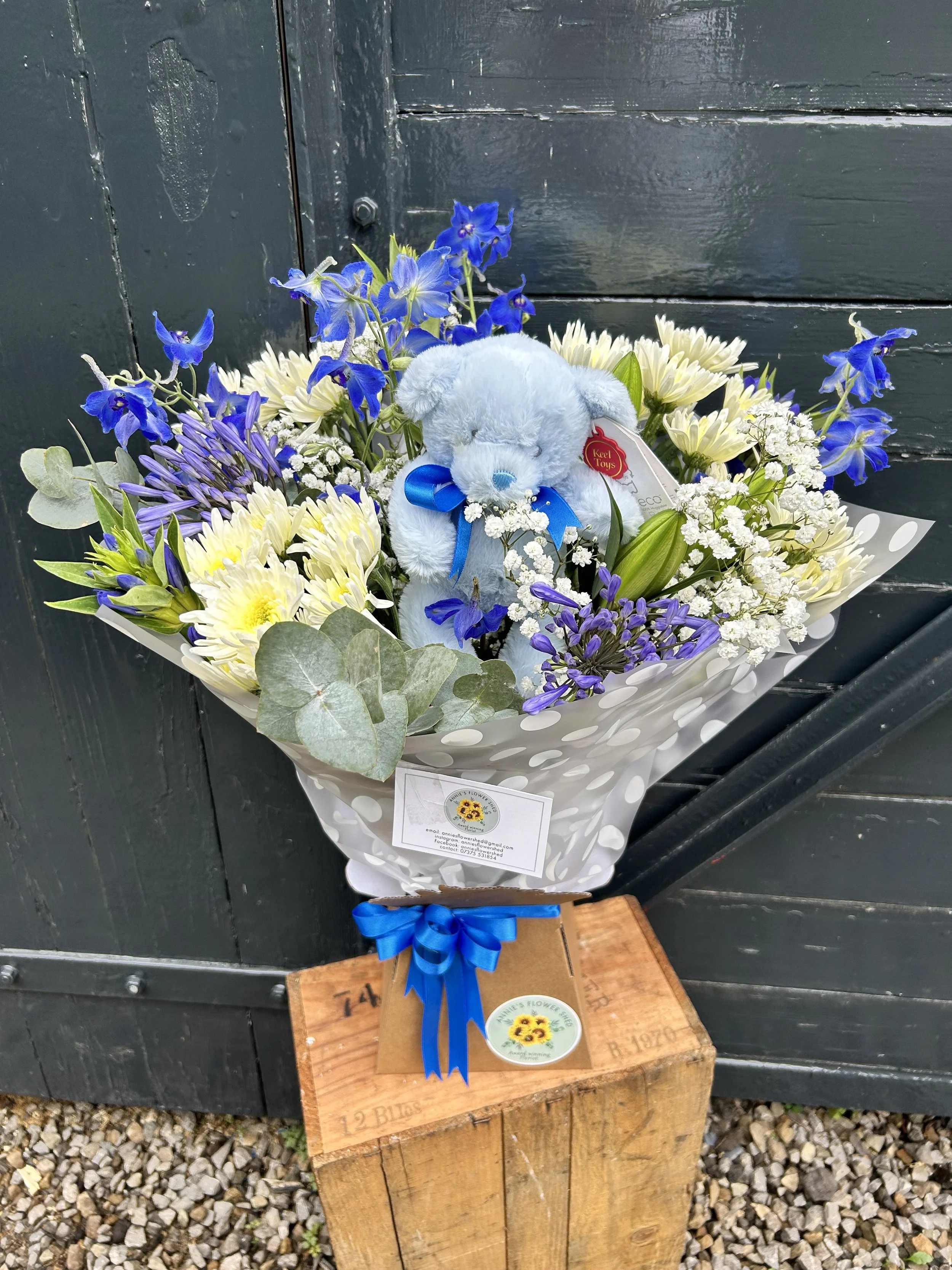 A bouquet of blue, white, and purple flowers with a gray plush teddy bear inside, adorned with a blue ribbon, placed on a wooden block against a dark wooden background.