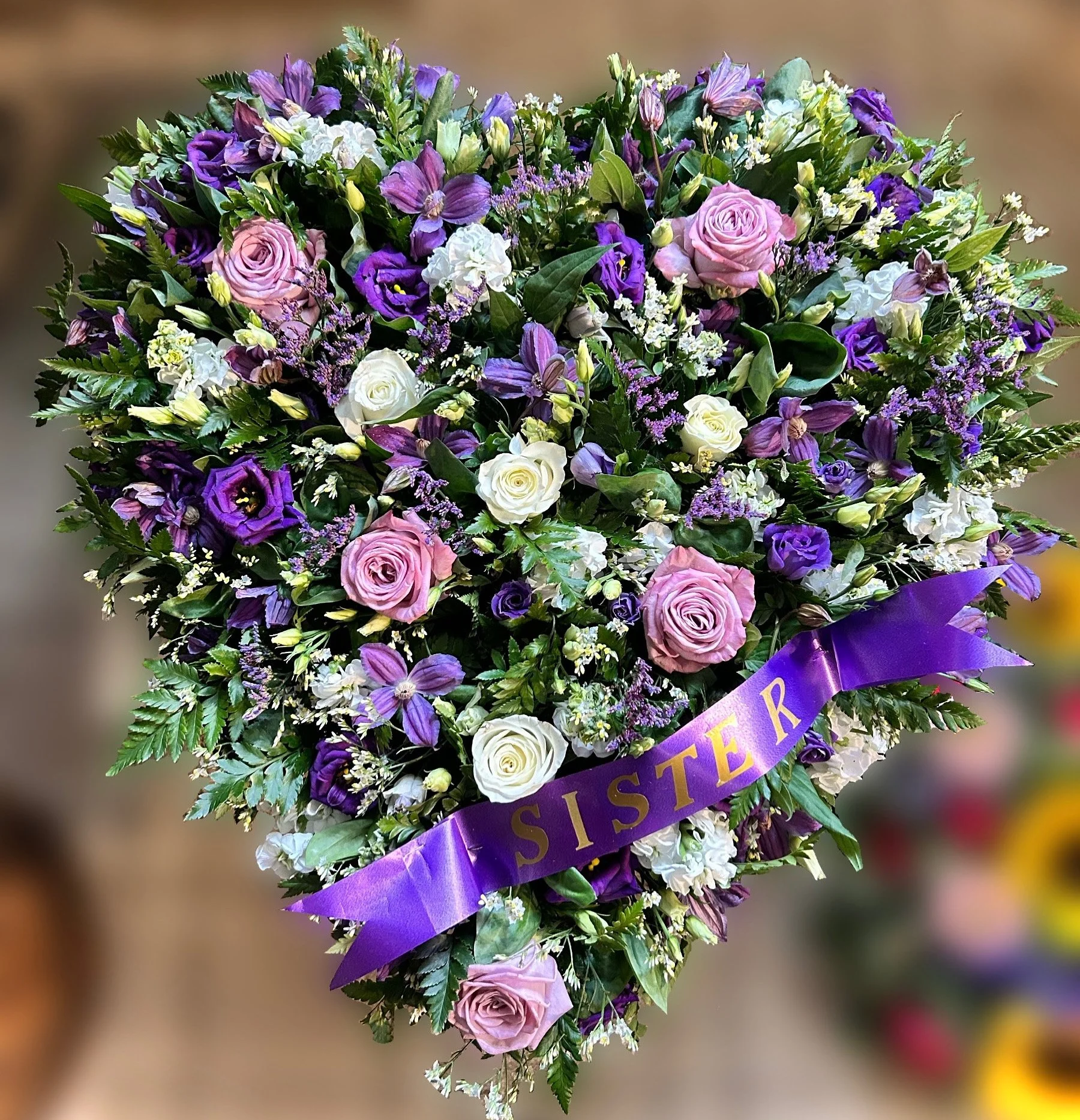 A floral arrangement featuring pink roses, purple lisianthus, and white flowers with greenery, decorated with a purple ribbon that reads 'SISTER'.