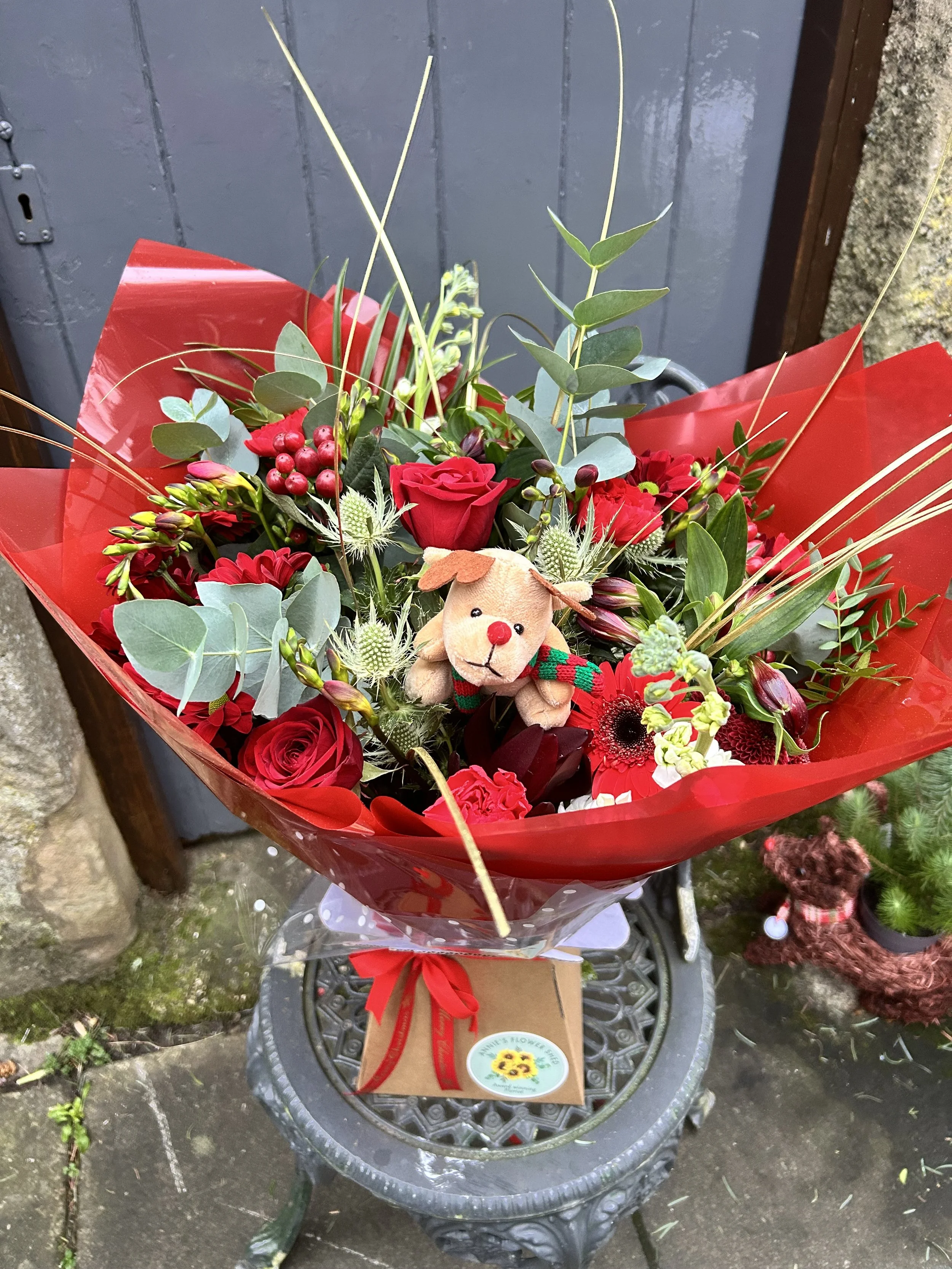 A bouquet of red and green flowers with a small stuffed reindeer figure, wrapped in red paper and attached to a gift box with a red ribbon, placed on an ornate metal table outside near a stone wall and grey door.