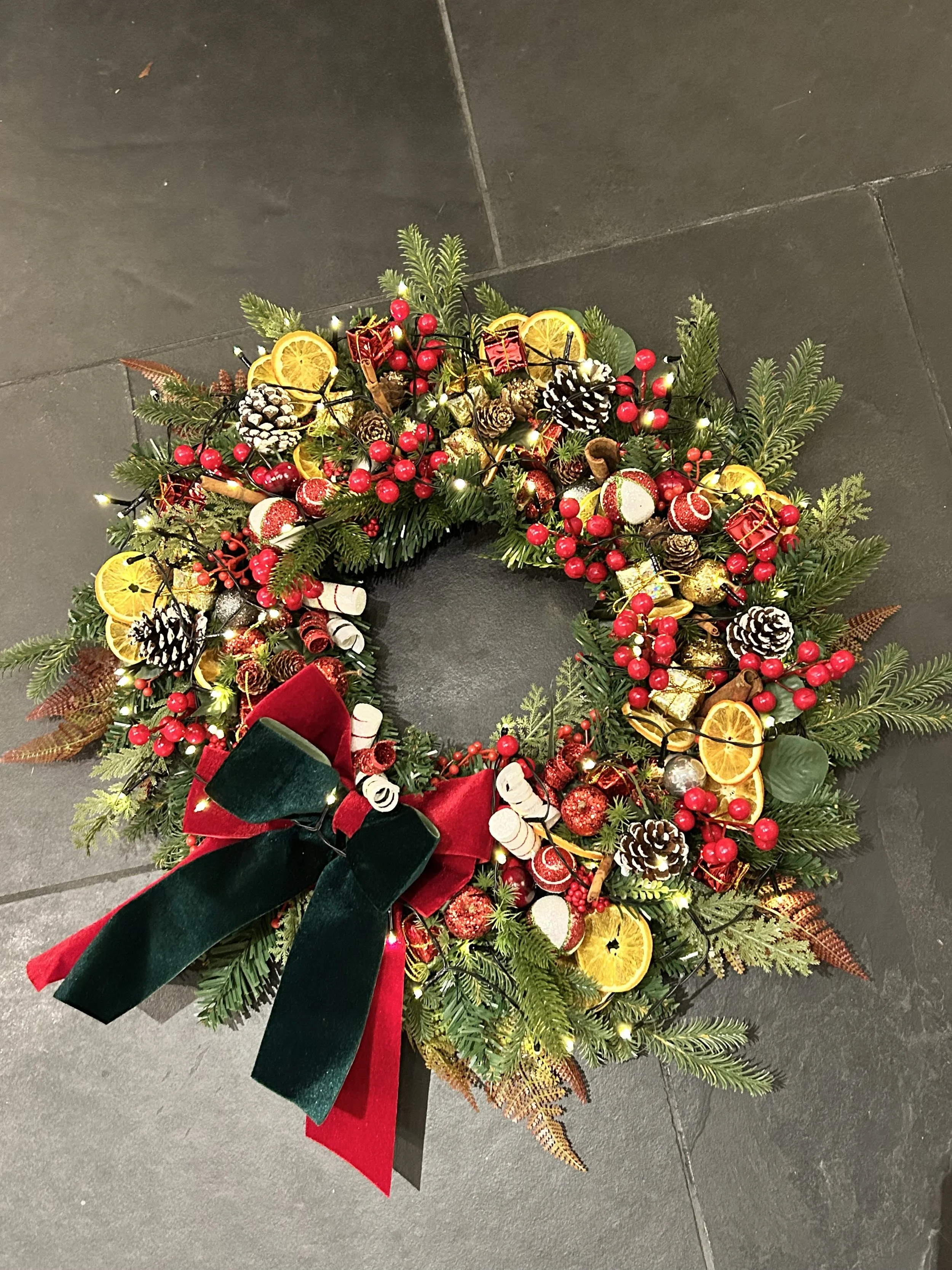 A Christmas wreath decorated with pine leaves, pinecones, red berries, dried orange slices, small wrapped gifts, and a large red and green velvet bow, illuminated with small string lights, on a dark tiled floor.