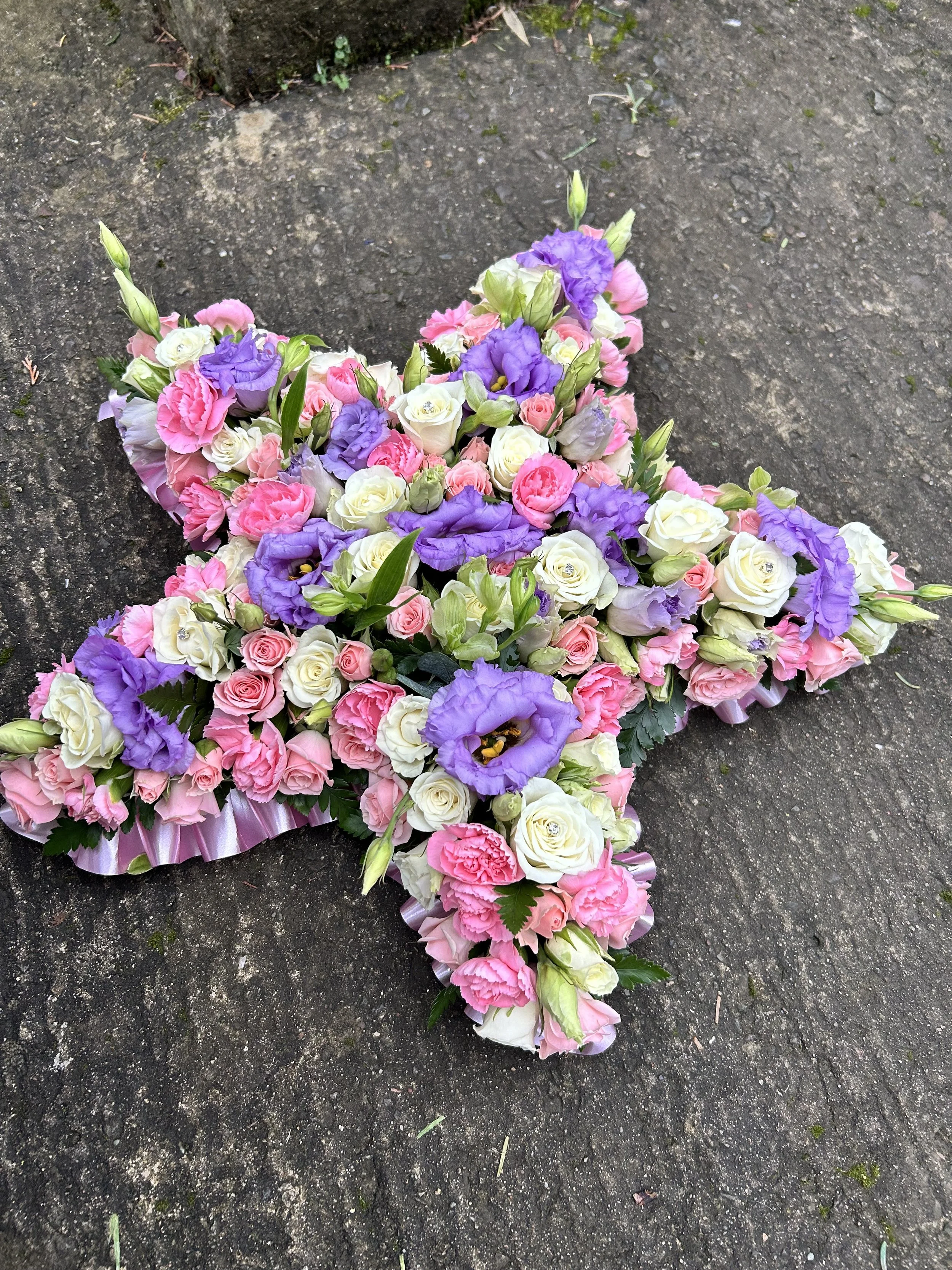 A large floral arrangement on the ground, featuring pink, white, and purple flowers, with some green leaves and buds, arranged in a cross shape.