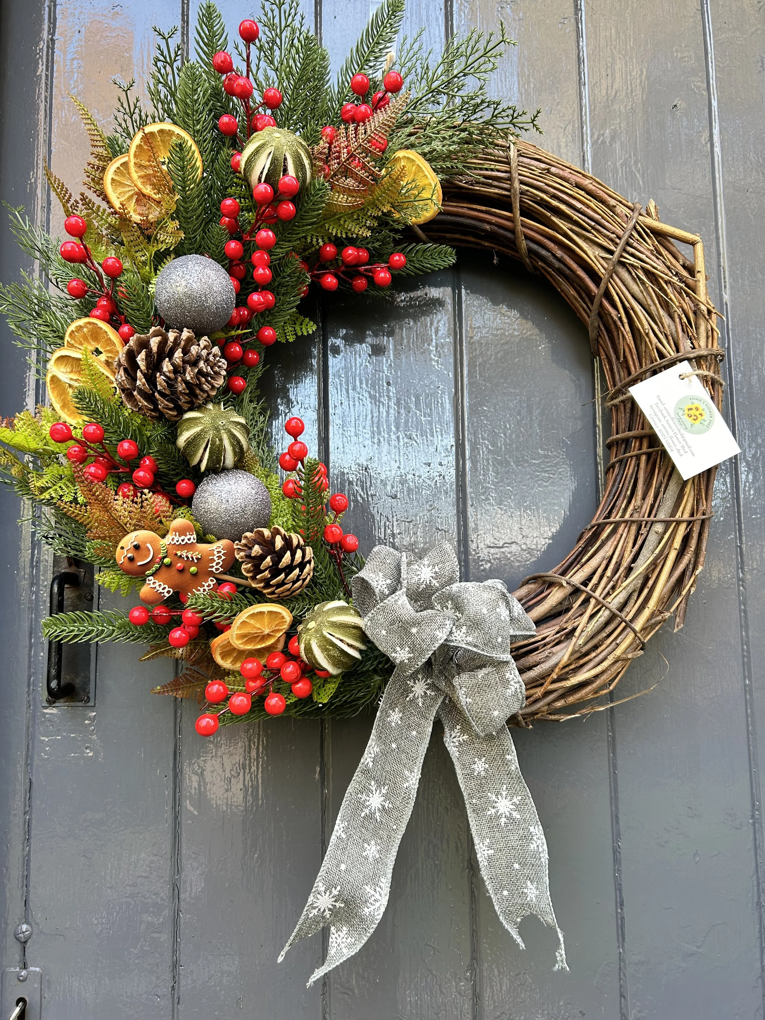 A Christmas wreath made of twisted natural branches decorated with pine branches, red berries, dried orange slices, pinecones, silver baubles, gold decorative elements, and a large silver bow, hanging on a blue wooden door.