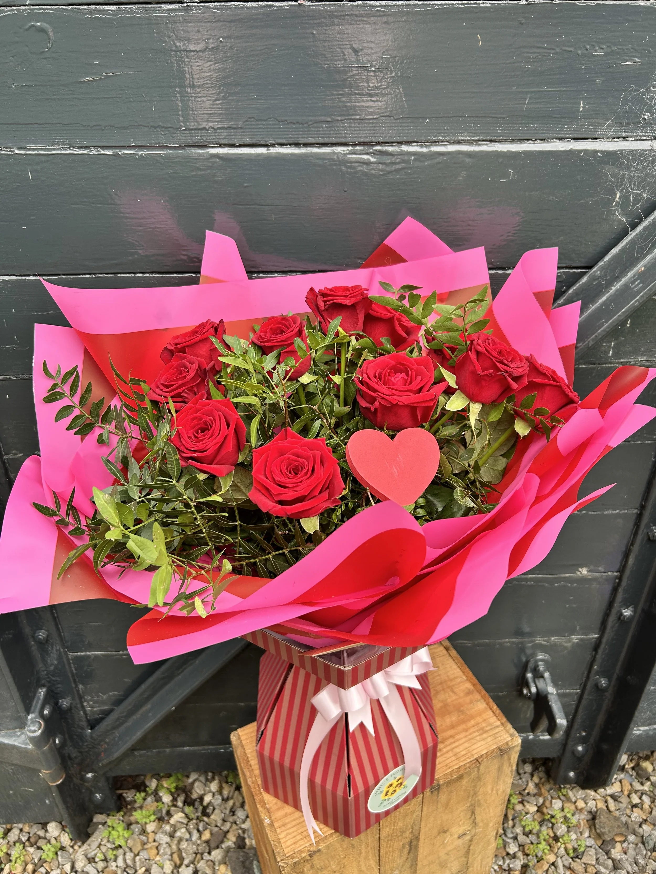A bouquet of red roses wrapped in pink and red wrapping paper with pink and white ribbons, placed on a wooden stand outside against a black wooden surface.