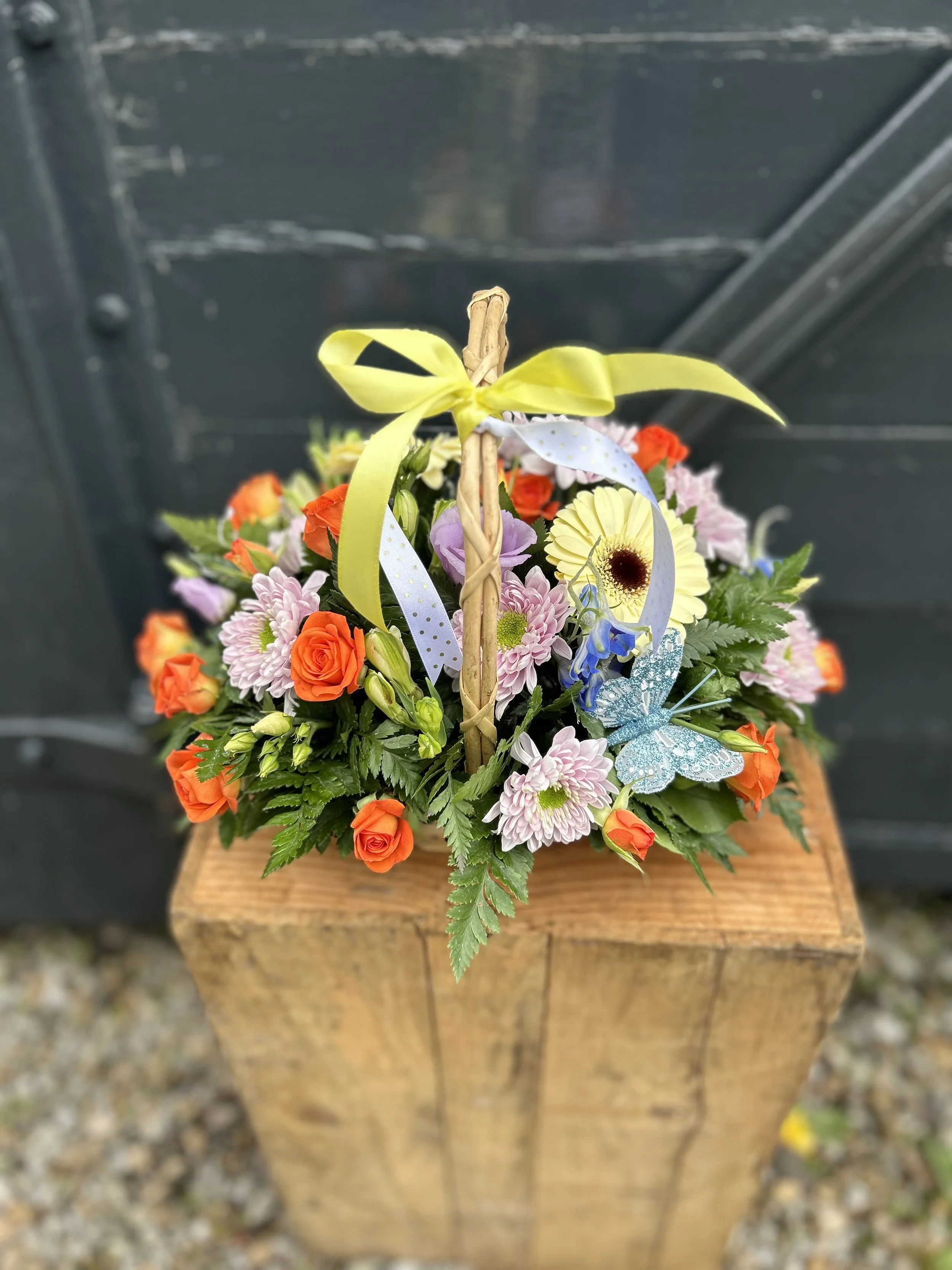 A colorful flower arrangement with pink, orange, yellow, purple, and blue flowers, decorated with a yellow ribbon and placed on a wooden surface.