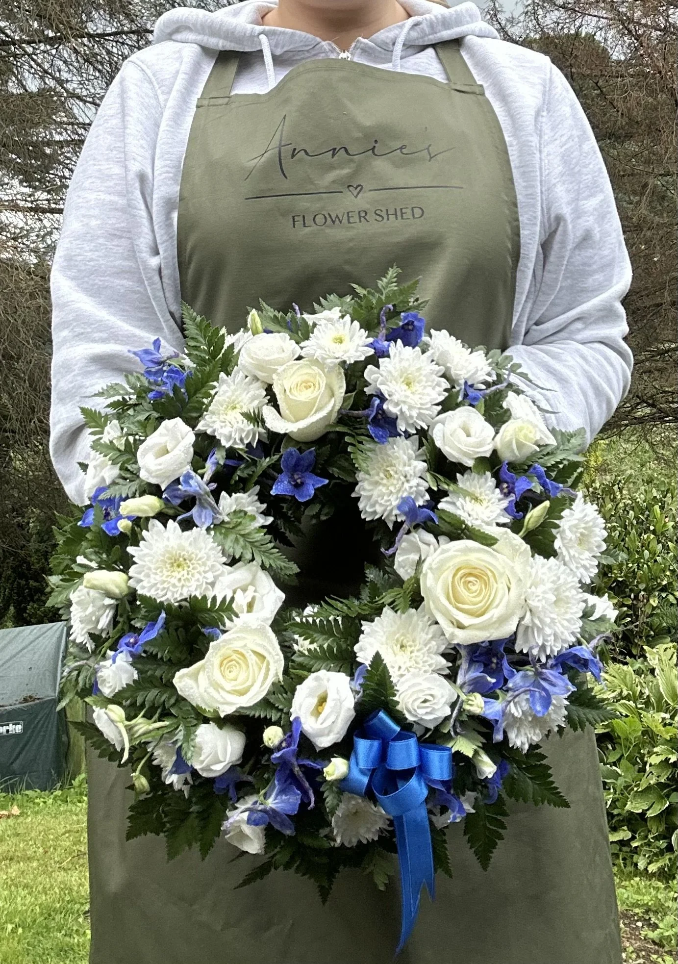 Person holding a large bouquet of white roses, white chrysanthemums, and blue flowers with a blue ribbon, standing outdoors in front of trees.