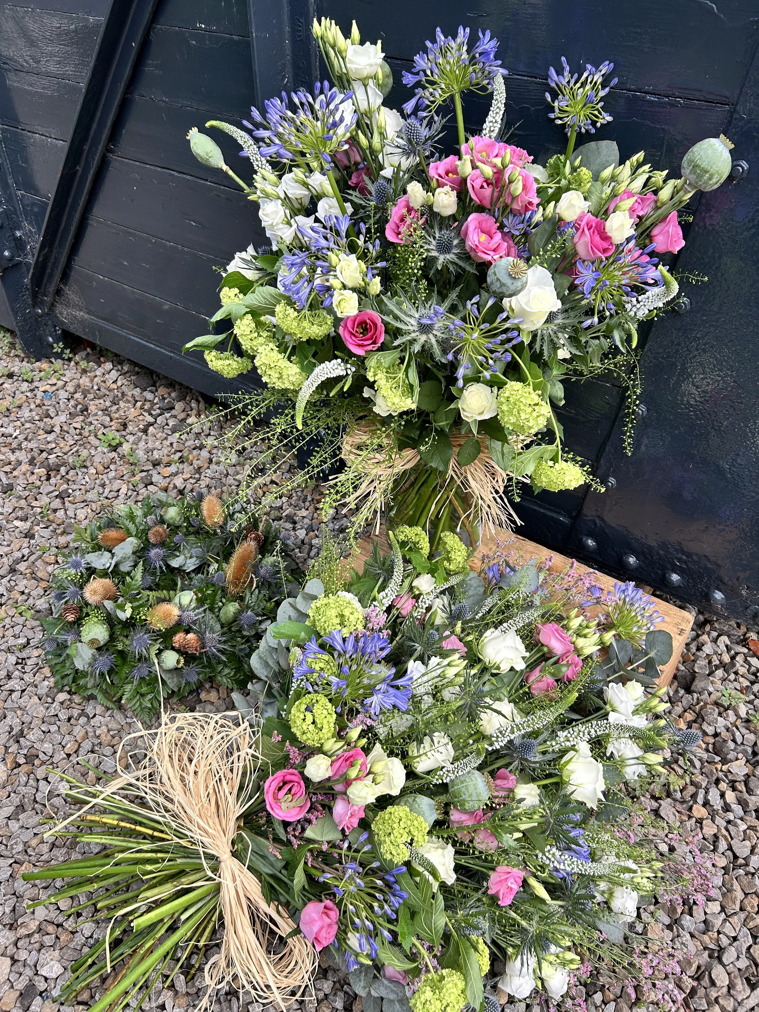 Vibrant floral arrangement with pink, white, and purple flowers on gravel ground, with a dark blue structure in the background.