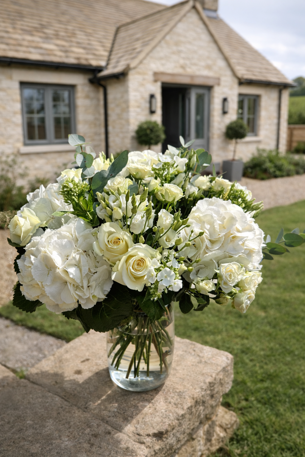 A bouquet of white roses, hydrangeas, and other flowers in a clear glass vase on a stone ledge outside a house with a brick exterior and well-kept garden.