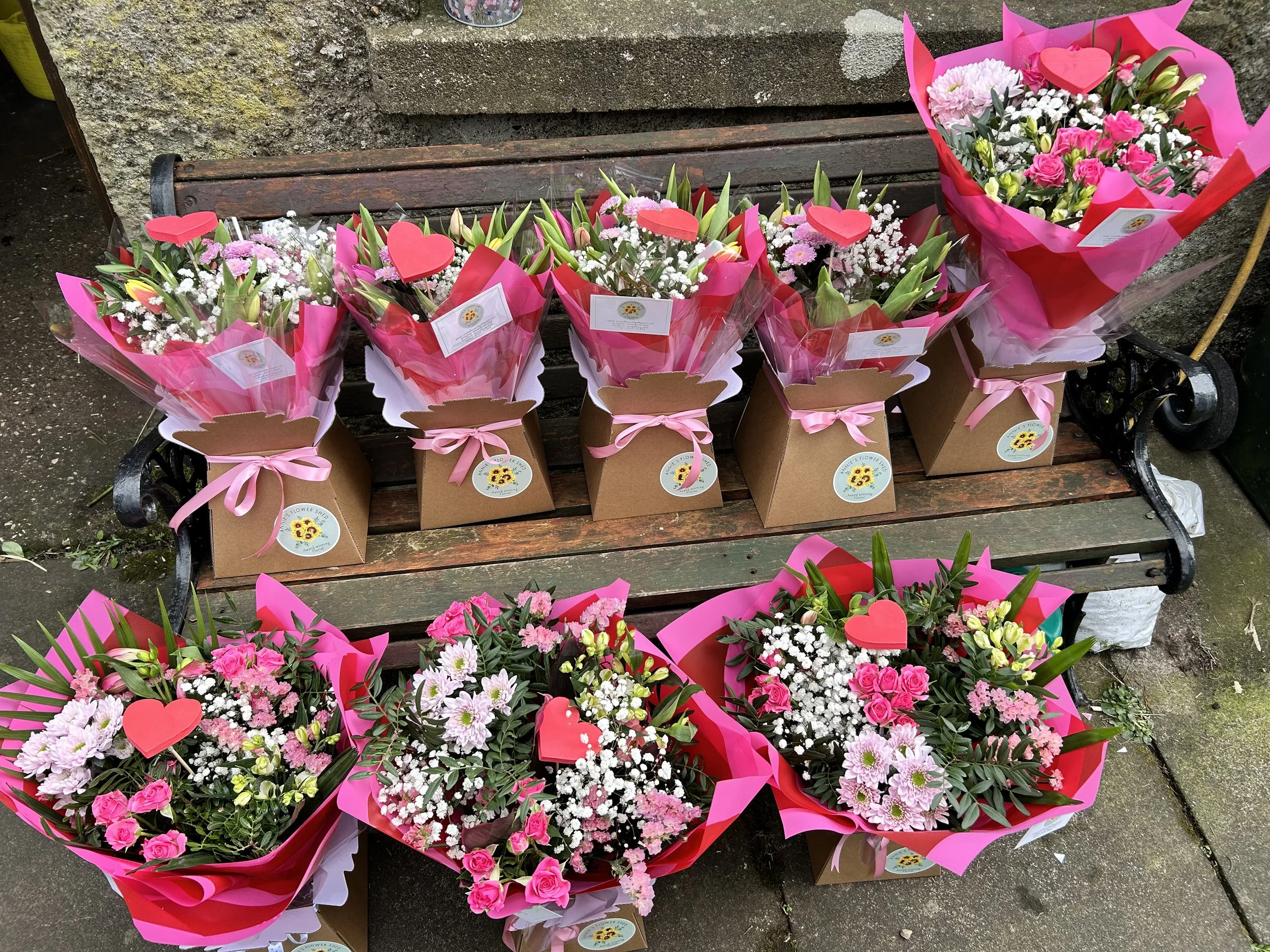 Multiple flower bouquets with pink and white flowers wrapped in pink paper, decorated with red heart-shaped toppers, placed on a wooden bench.