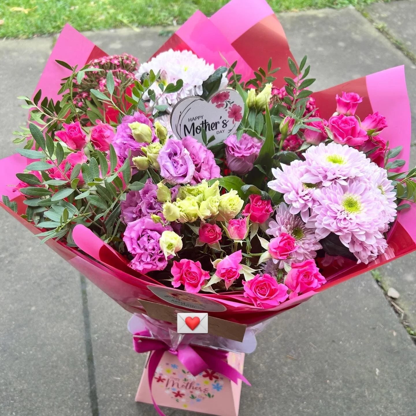 A colorful Mother's Day bouquet with pink, purple, and white flowers, including roses and chrysanthemums, wrapped in pink paper with a small card and decoration.