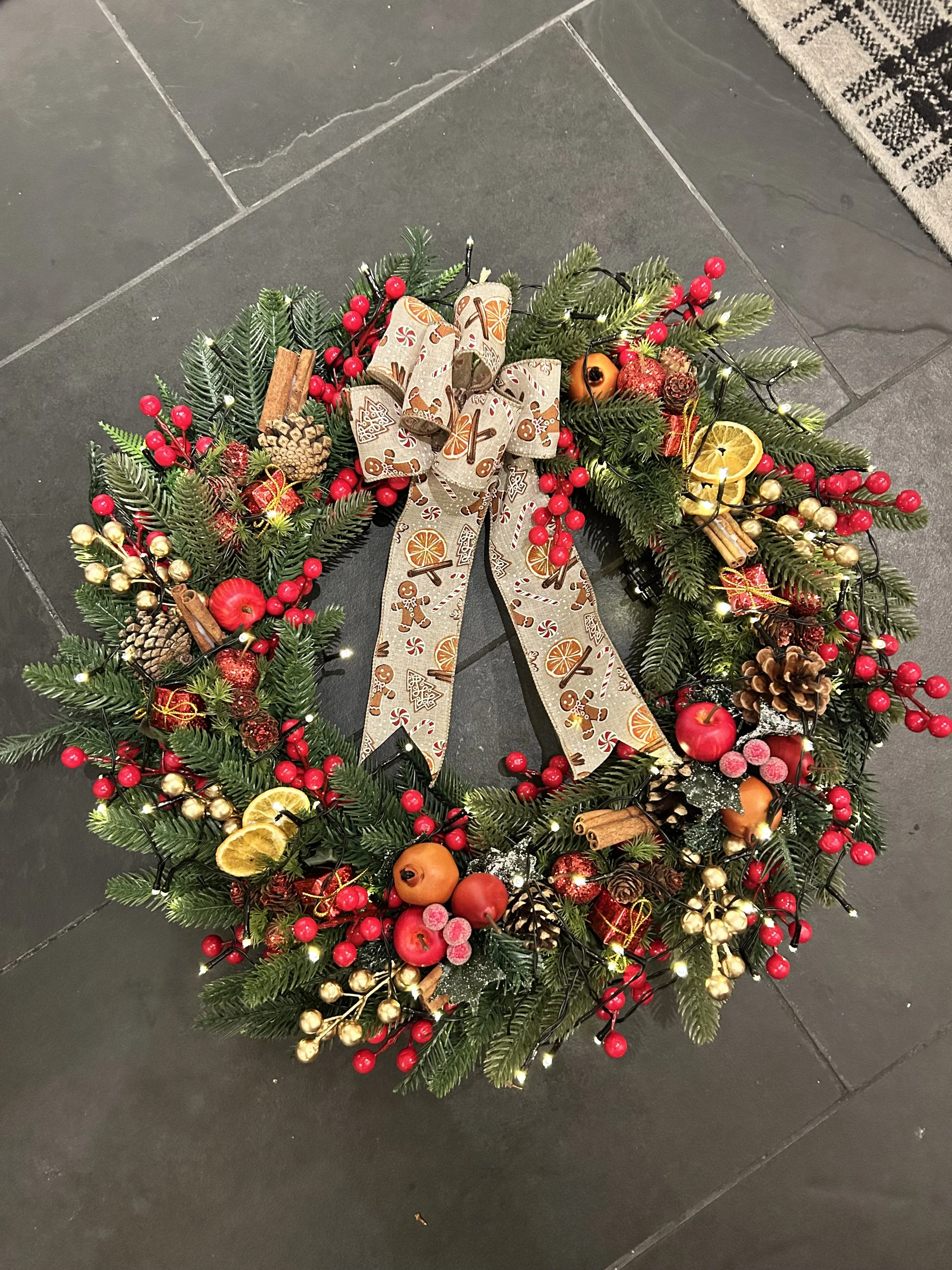 A Christmas wreath decorated with red berries, pine cones, dried orange slices, cinnamon sticks, small wrapped presents, and a bow with holiday patterns, placed on a dark tiled floor.