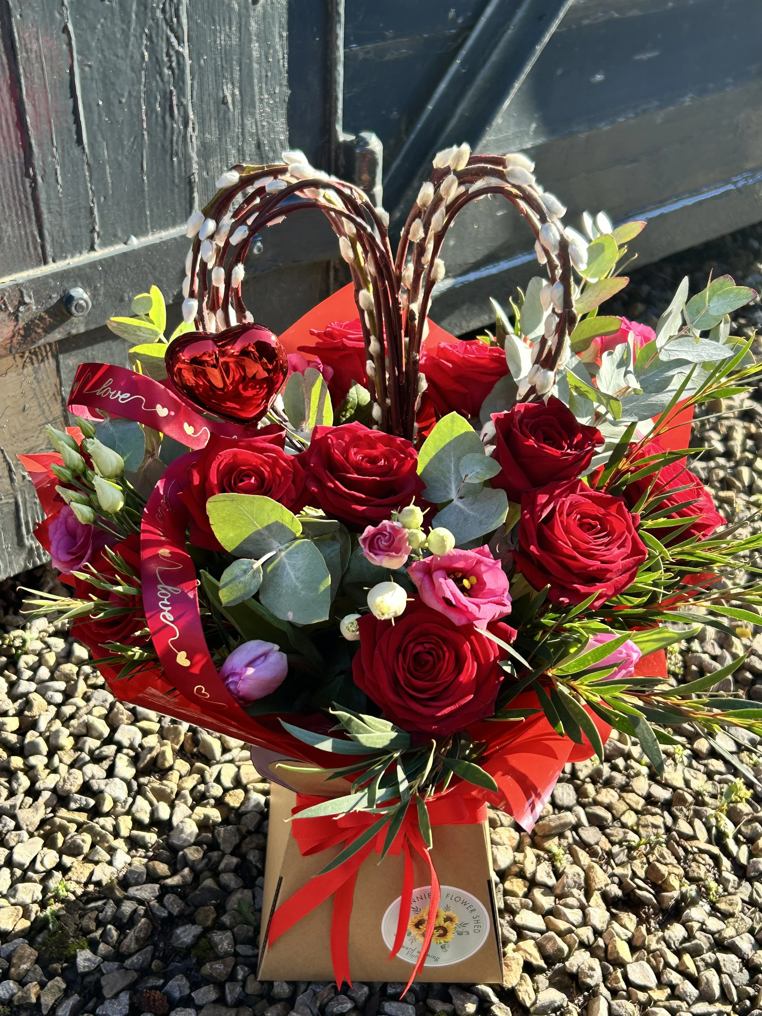 A flower arrangement with red roses, pink flowers, greenery, and a red heart-shaped ornament in a basket, with a ribbon that has hearts and the word "Love" on it, placed on top of a cardboard box on a gravel surface outdoors.