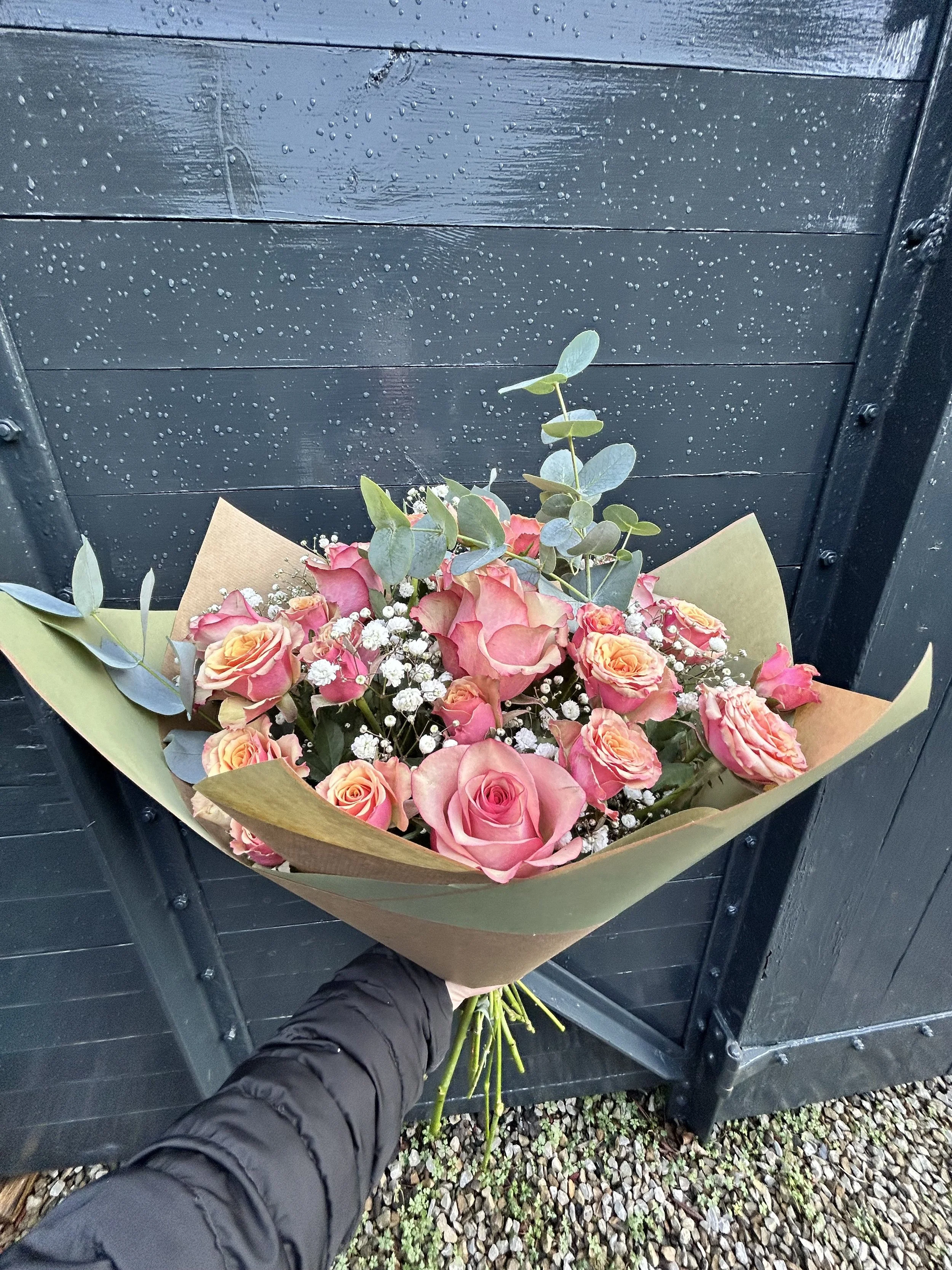 A bouquet of pink and peach roses, white baby's breath, and green leaves in wrapping paper, being held by a person wearing a black jacket, in front of a black metal fence with raindrops on it.