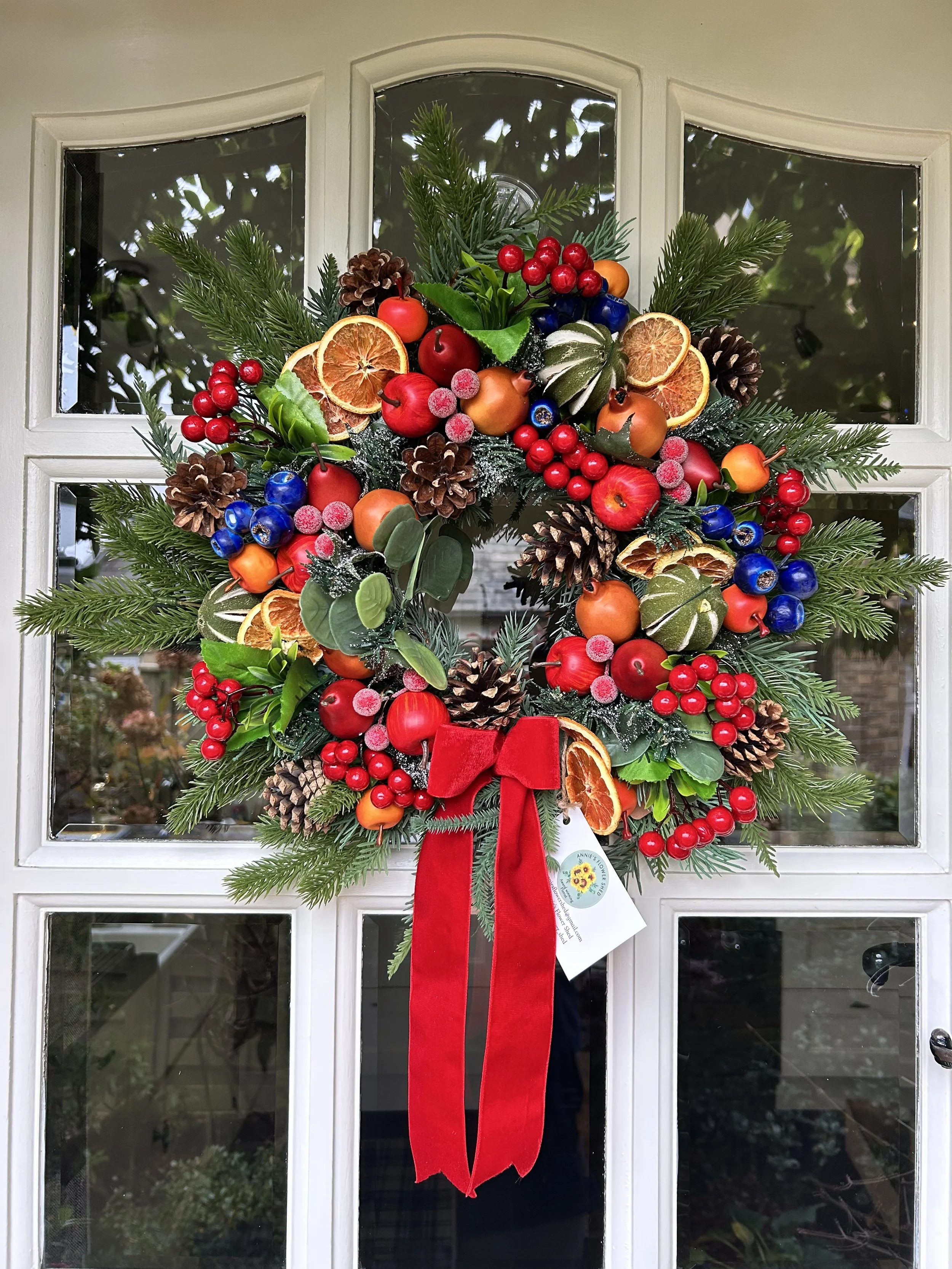A Christmas wreath decorated with dried orange slices, red apples, pinecones, red berries, green foliage, and a red bow is hanging on a white door with glass panels.