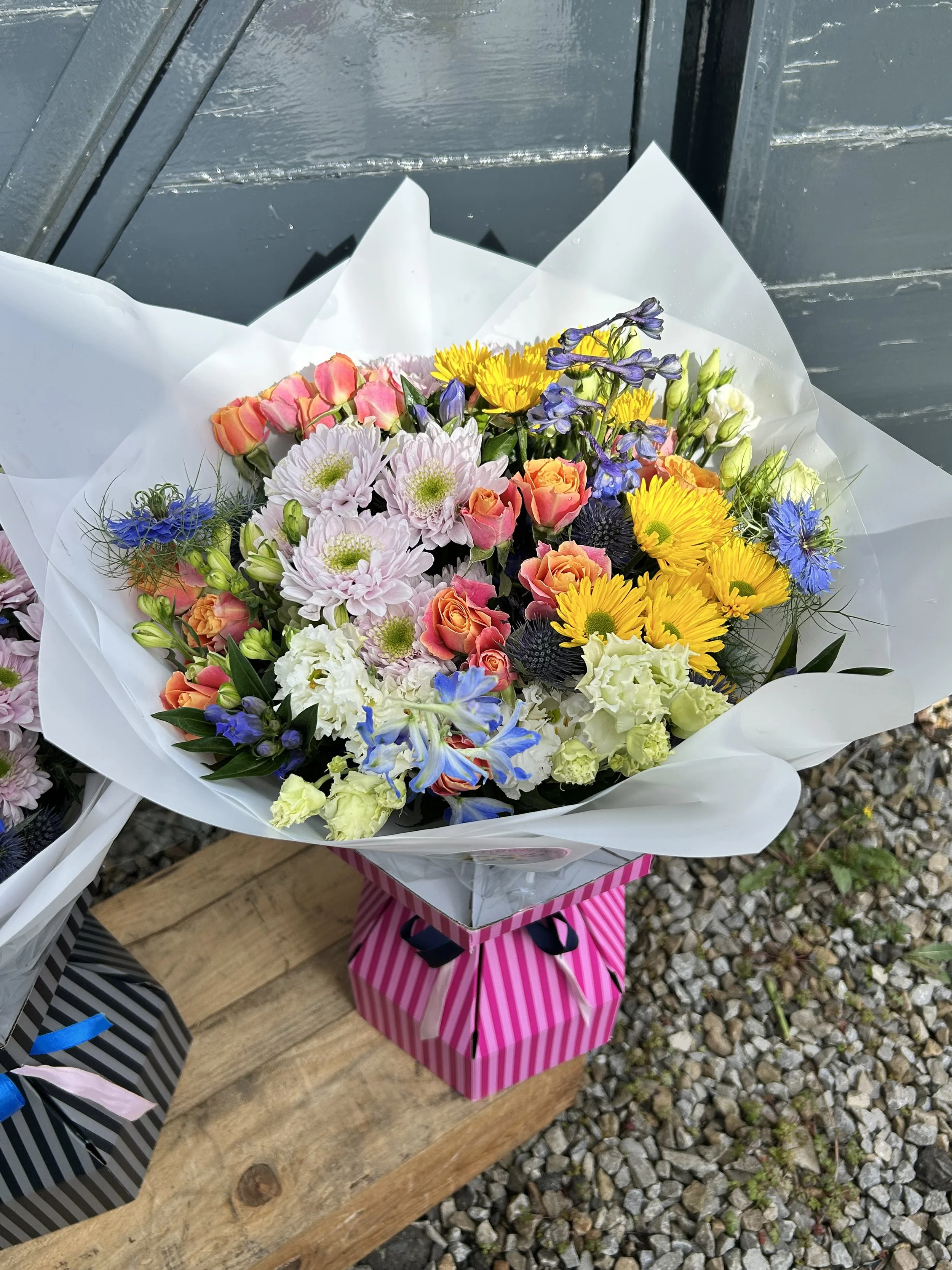 A colorful bouquet of flowers in white wrapping paper, placed in a pink and white striped gift bag, on a wooden surface with a gravel background.