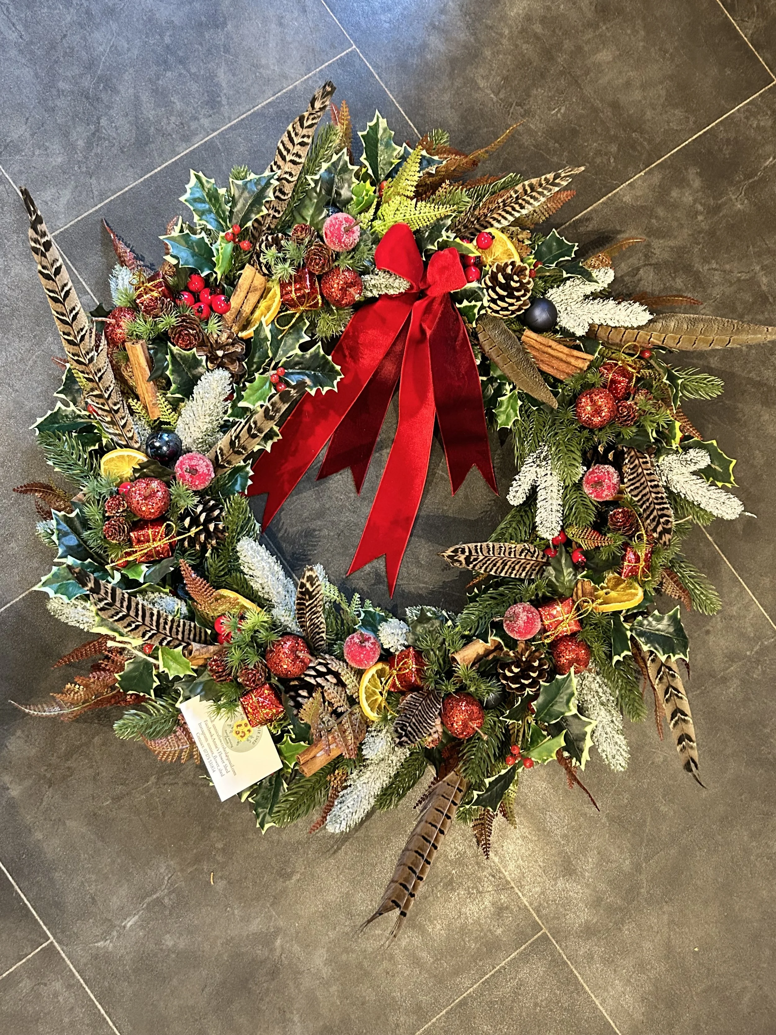 A Christmas wreath decorated with holly leaves, red berries, pine cones, dried orange slices, cinnamon sticks, glittery red ornaments, gold ribbons, white frosted branches, and large feathers with black and white stripes.
