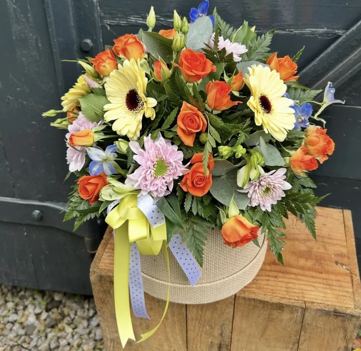 A bouquet of mixed flowers including roses, daisies, and other blooms in a round beige basket with a yellow and blue polka dot ribbon, placed on a wooden block.