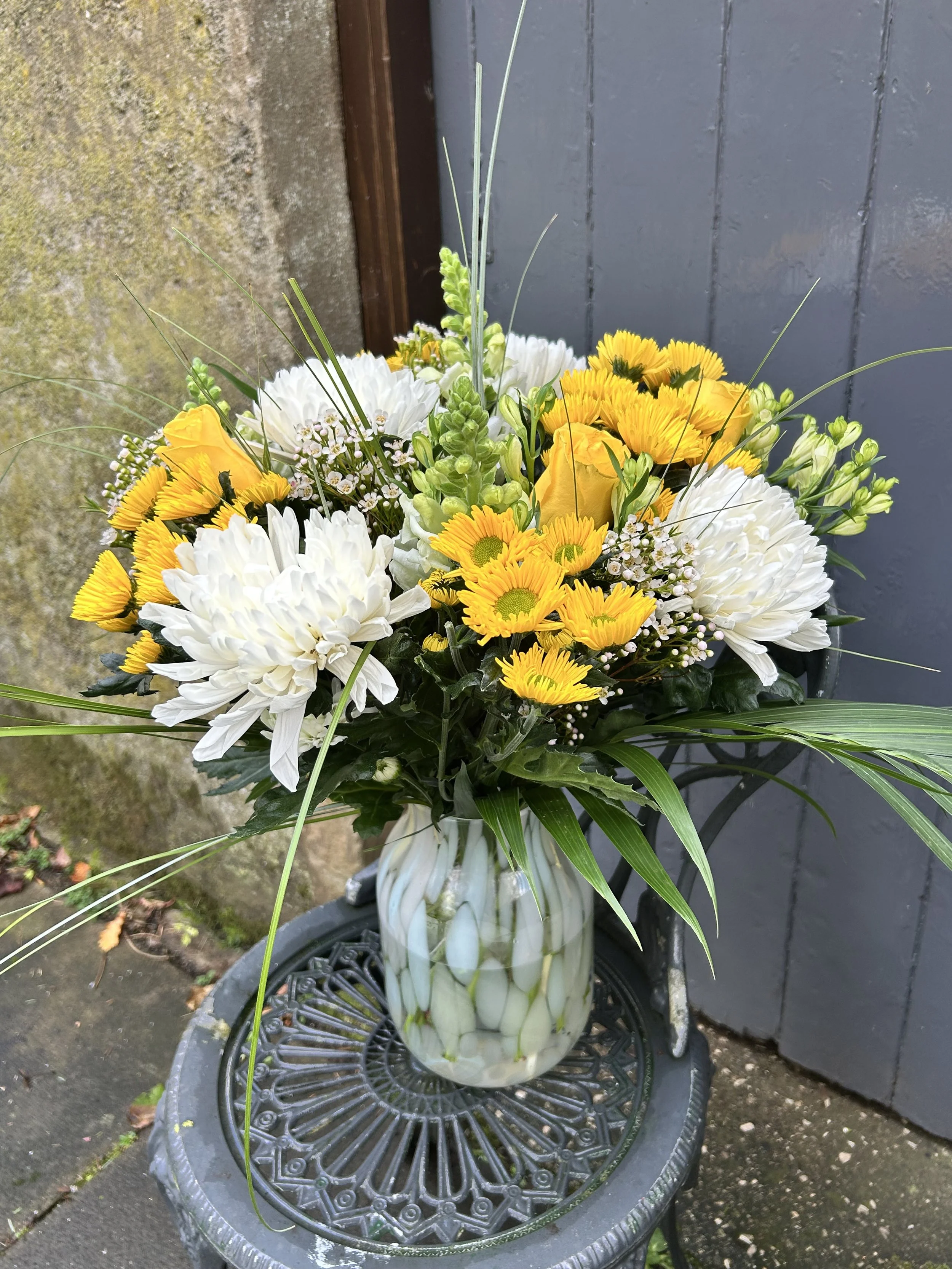 A floral arrangement in a glass vase placed on a decorative black metal table outdoors, with a dark gray wooden wall and mossy ground in the background.