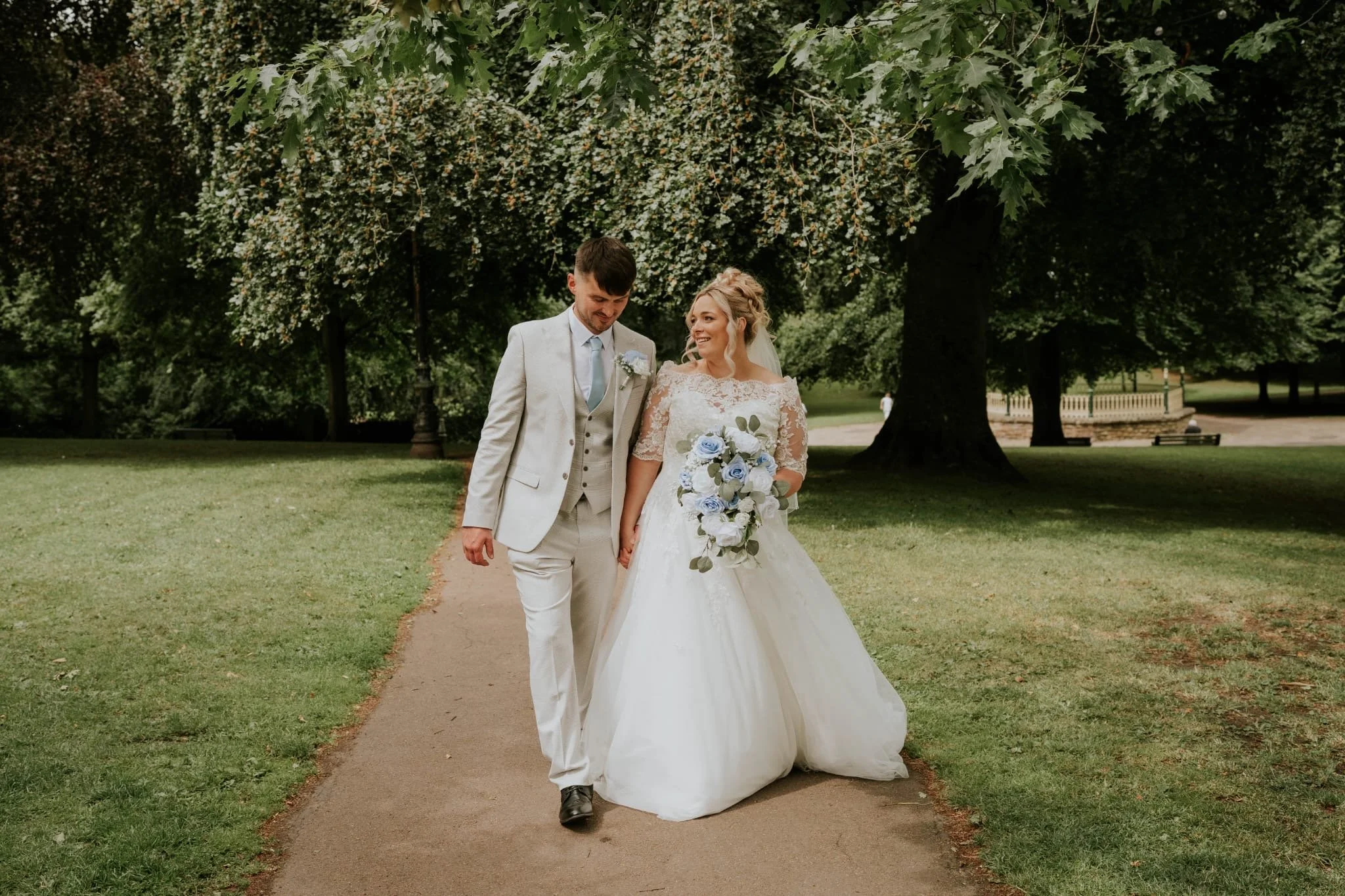 A bride and groom walking together outdoors in a park, with the bride holding a bouquet of blue and white flowers, smiling and looking at the groom. The groom is dressed in a light suit and tie, and they are walking along a dirt path surrounded by gr