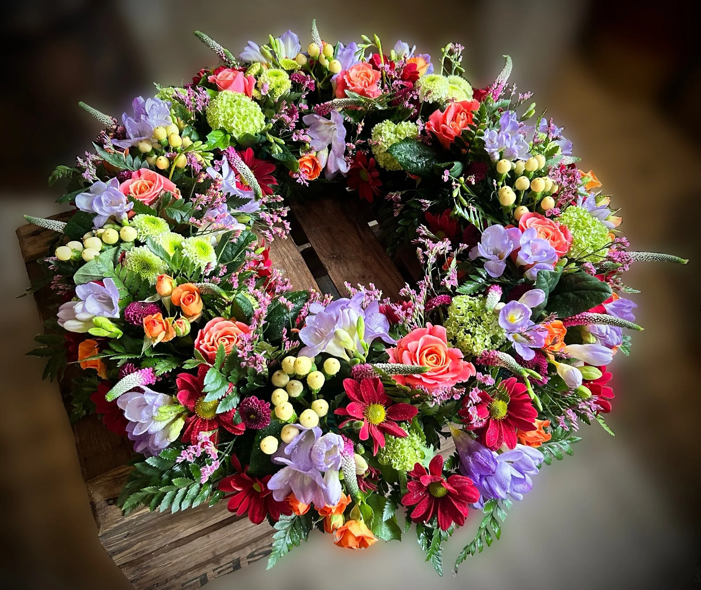Colorful flower arrangement featuring pink roses, purple and green hydrangeas, red daisies, white and purple lisianthus, yellow berries, and surrounding greenery on a wooden surface.