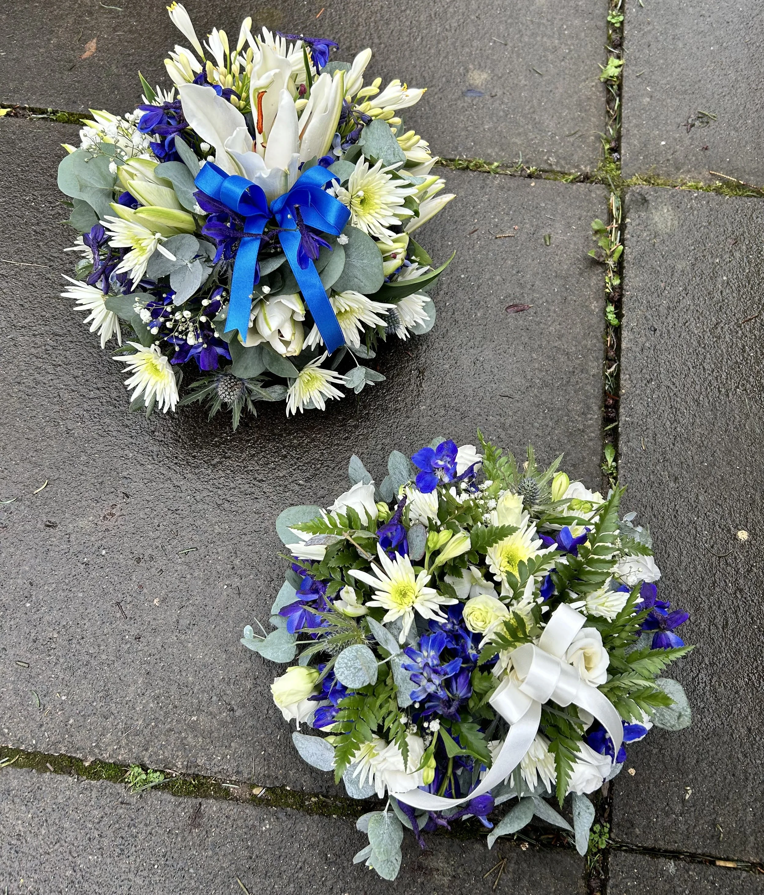Funeral flower arrangement with white and blue flowers, green leaves, and a blue ribbon, placed on a concrete sidewalk.