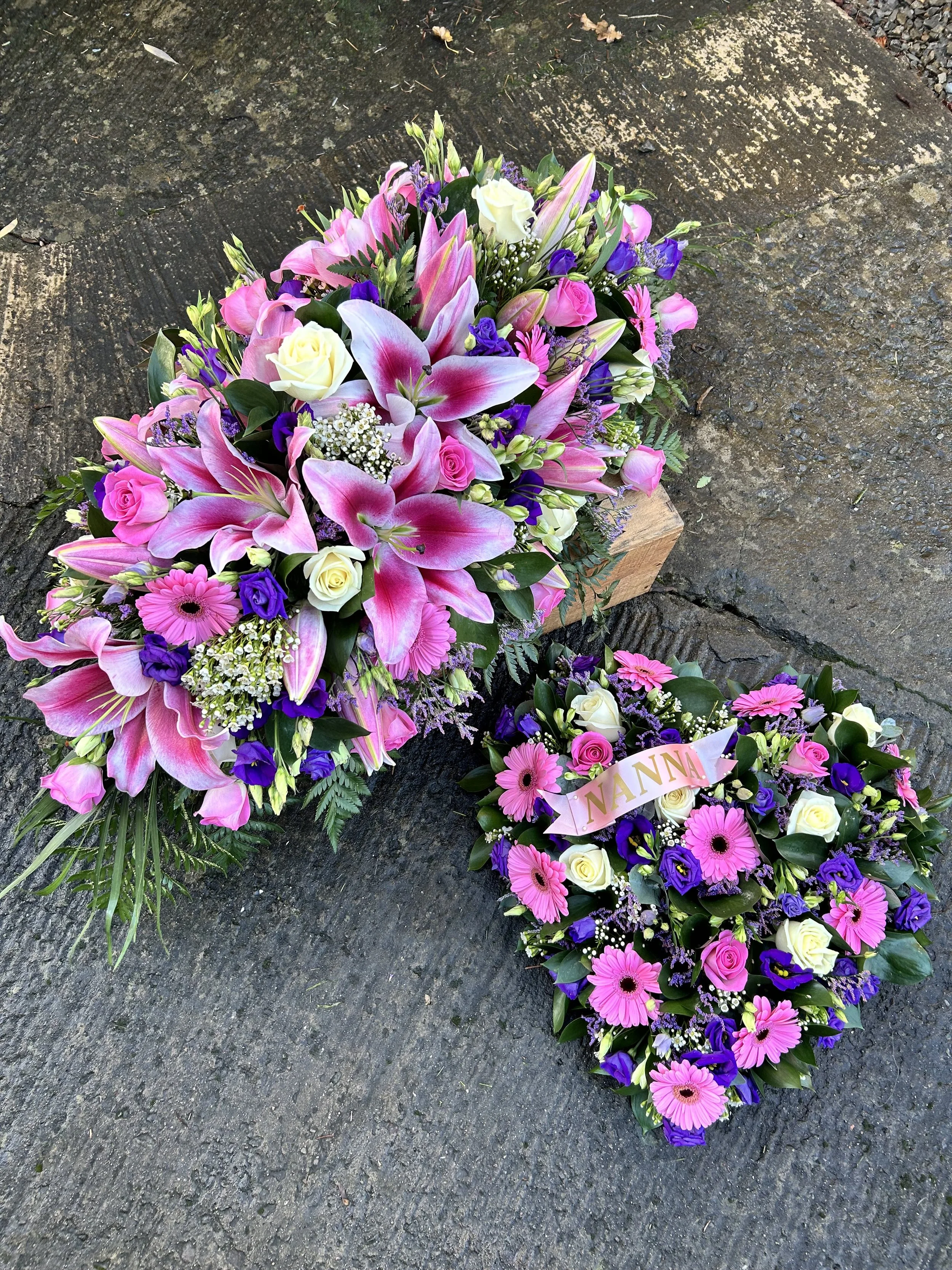 A floral casket spray with pink lilies, white roses, purple lisianthus, pink gerbera daisies, and assorted greenery, resting on a rough concrete surface.