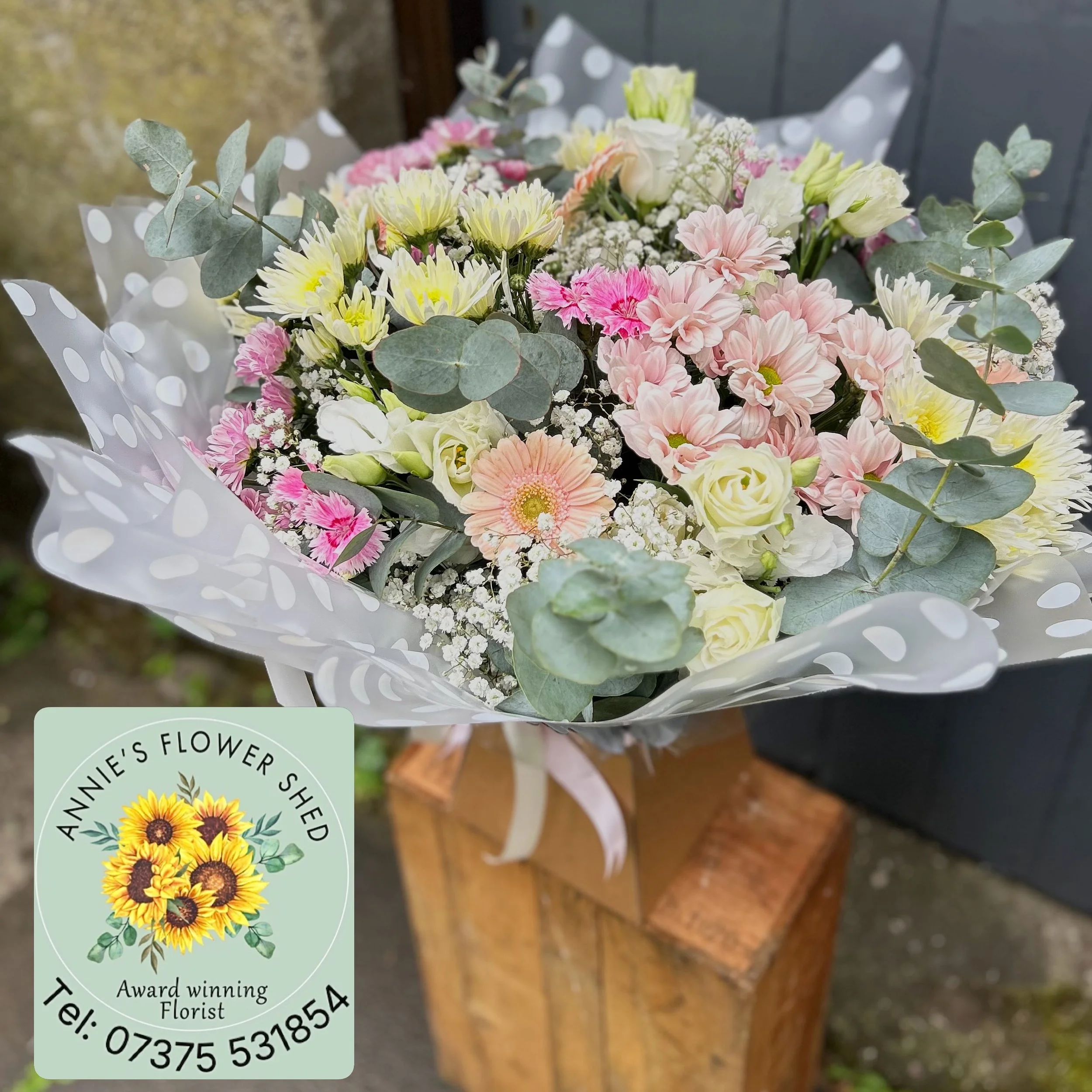 A bouquet of pink, yellow, white, and peach flowers with greenery and baby's breath wrapped in white polka-dotted paper.