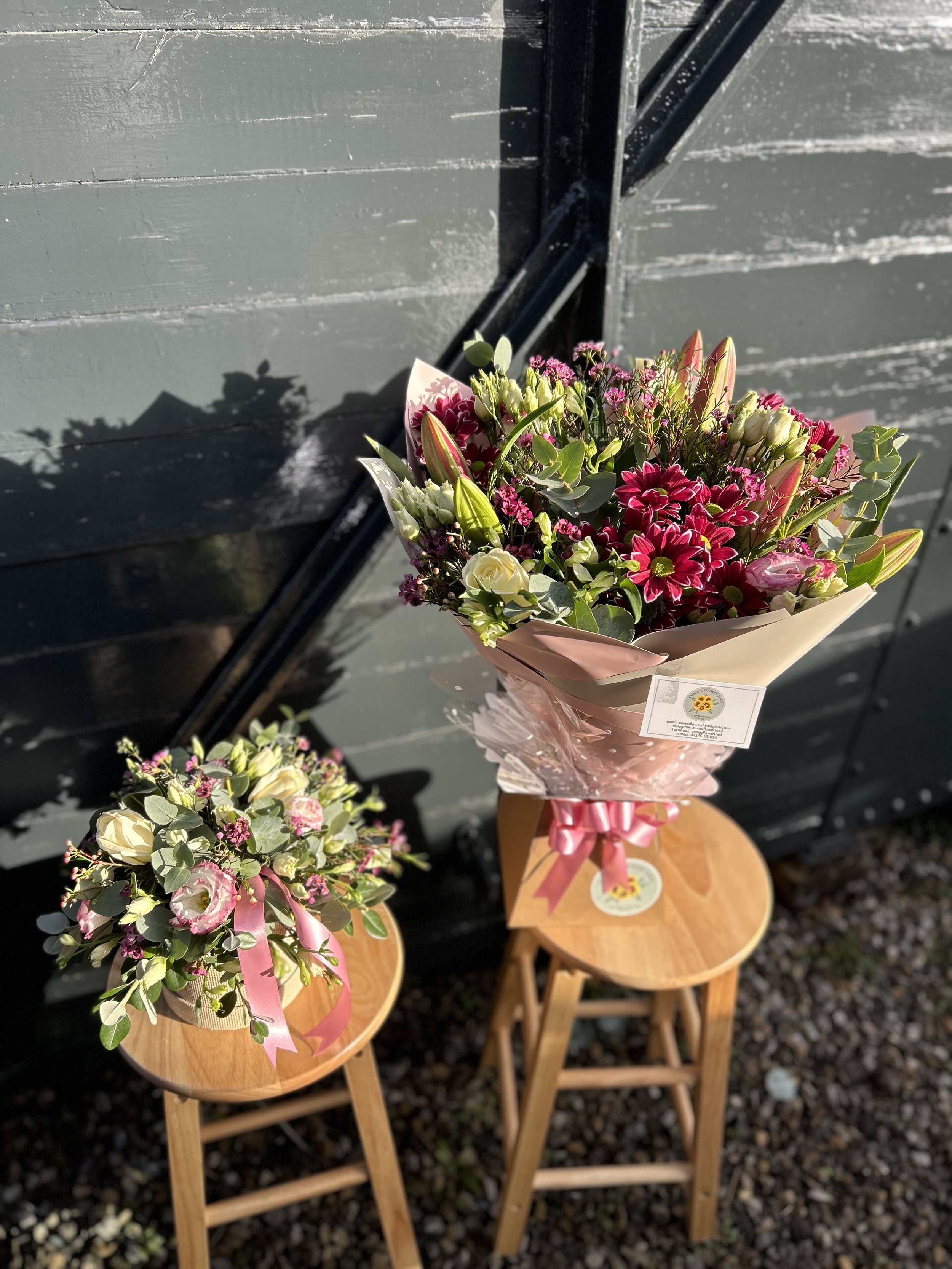 Two wooden stools each holding a bouquet of flowers, one with pink and white flowers and the other with mainly white flowers, against a dark gray wall with shadows cast by the sunlight.